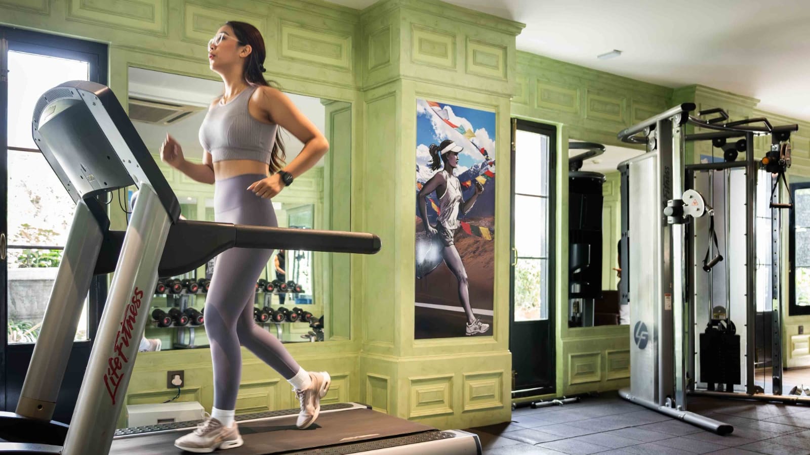 A guest running on a treadmill inside the Vyayam Fitness Centre at Nepali Ghar Hotel, framed by sage-green heritage panelling and a Himalayan running mural.