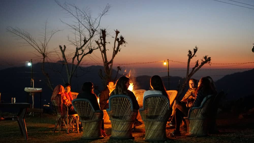 Guests in wicker chairs gathered round a bonfire on the lawn at dusk, the hills behind