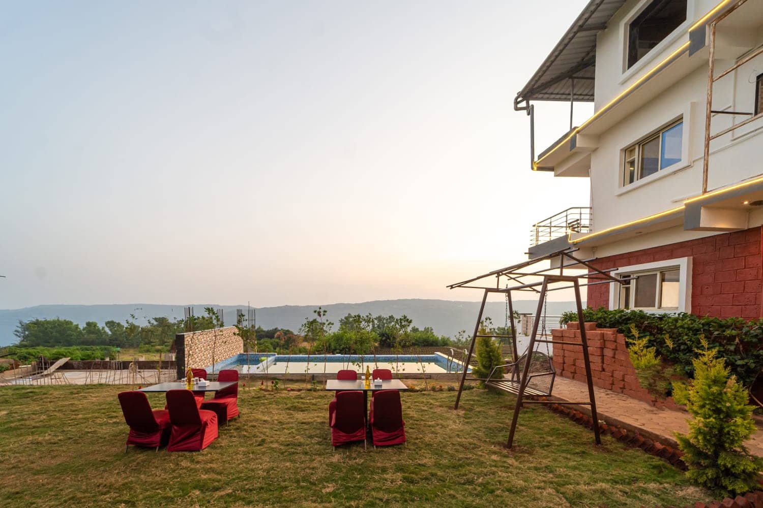 Lawn with red-cushioned tables, a garden swing and the pool beside the resort at dusk