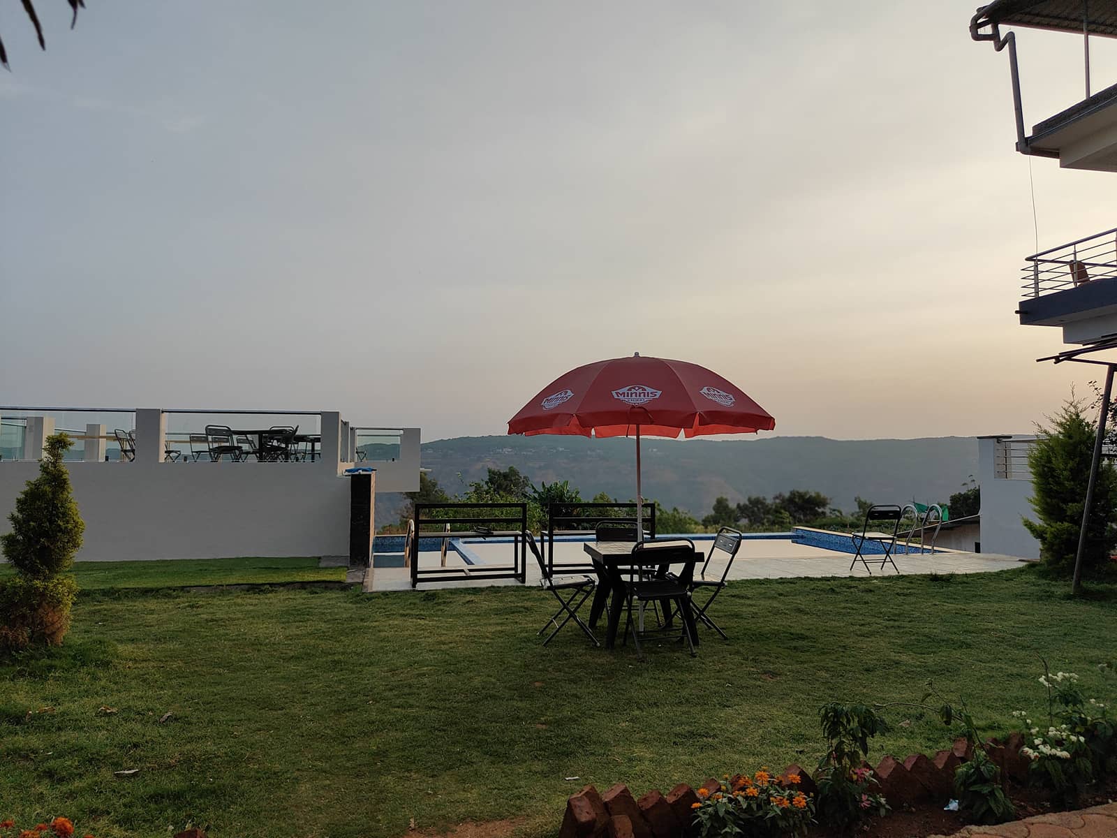 A table under a red parasol on the lawn, with the pool and hills behind at dusk