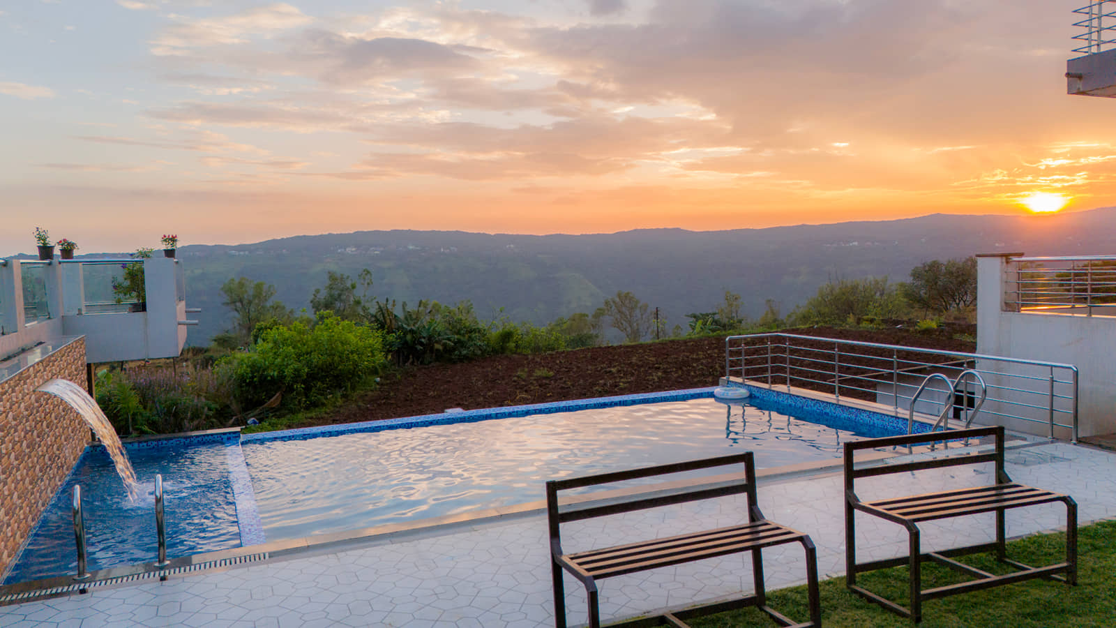 Infinity swimming pool with a waterfall spout and wooden benches, the valley glowing at sunset beyond