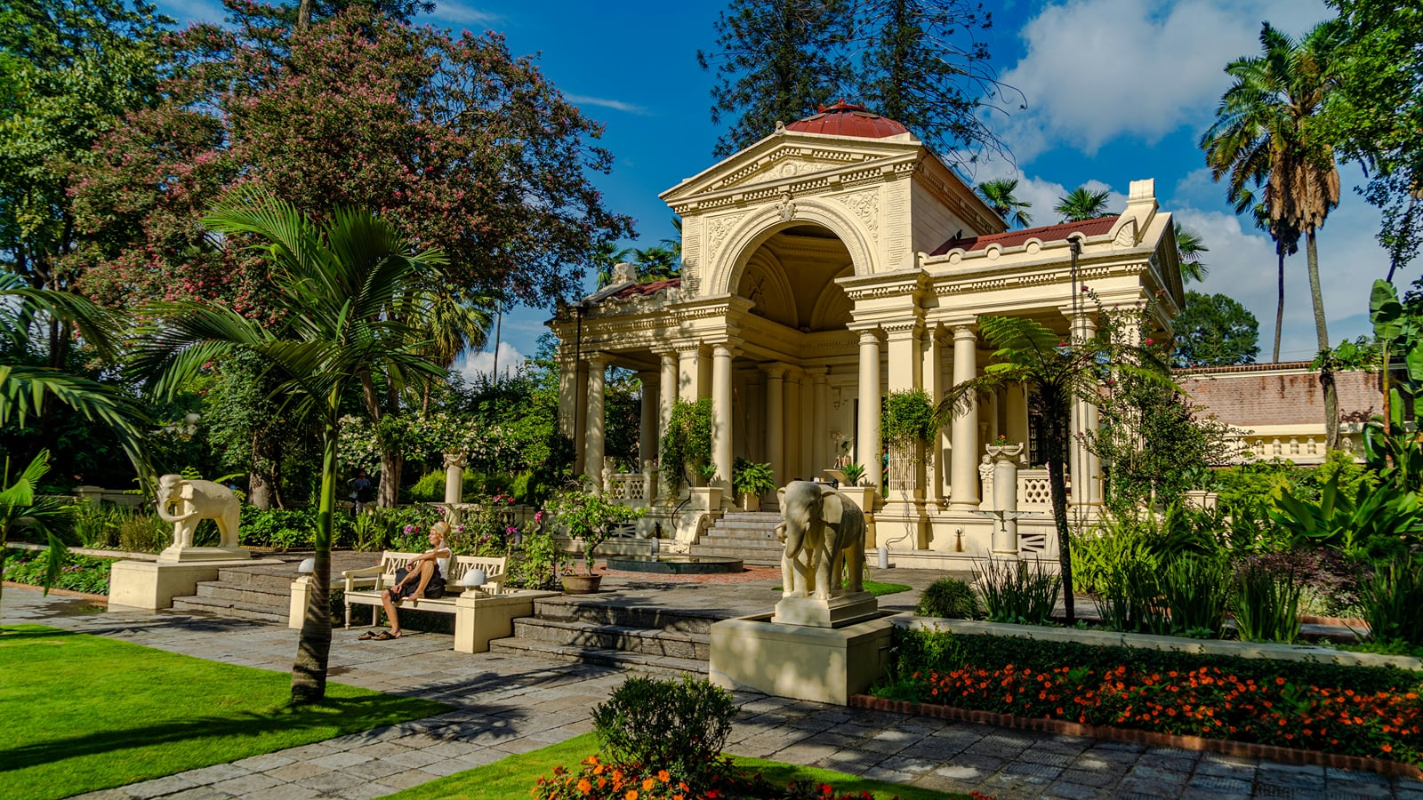 The Garden of Dreams Kathmandu, a Neo-Classical pavilion with an arched portico and stone lion statues, set among palms and flowerbeds.