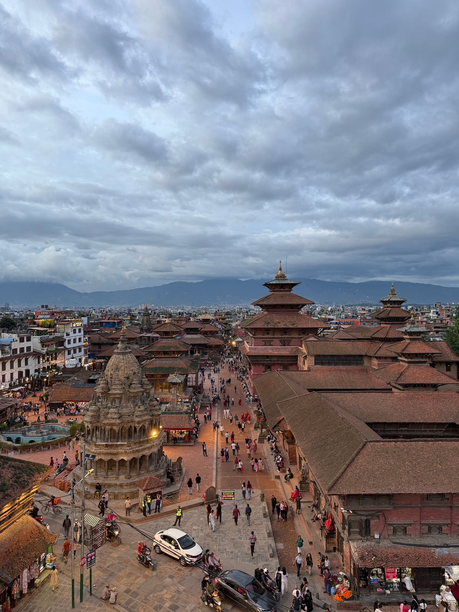 An aerial view of Kathmandu Durbar Square at dusk, with its stone shikhara temple, tiered red-brick pagodas, and visitors walking the paved square beneath a cloudy sky.