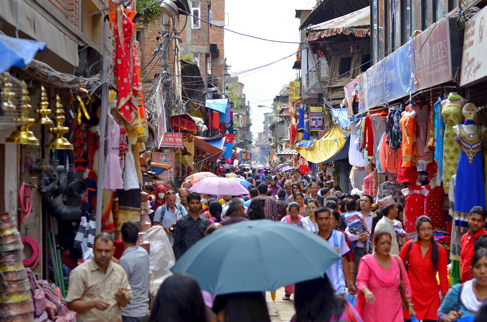 The Thamel market near Thamel Chowk in Kathmandu, a crowded lane for shopping in Thamel, with shoppers beneath umbrellas and stalls hung with colourful clothing, brass bells, and handicrafts.