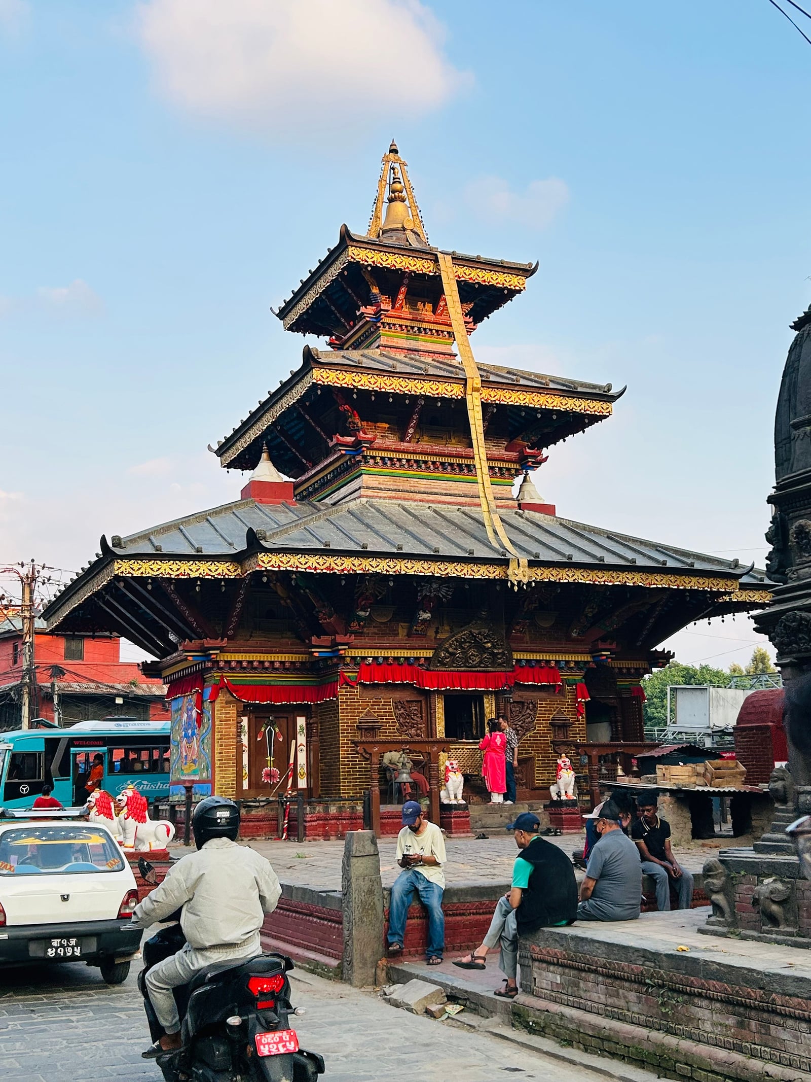 Bhagwan Bahal, a Buddhist monastery courtyard off a Thamel street in Kathmandu, with a gilded, tiered Newari pagoda, a yellow ceremonial banner hanging from its roof, and visitors resting on the stone steps.