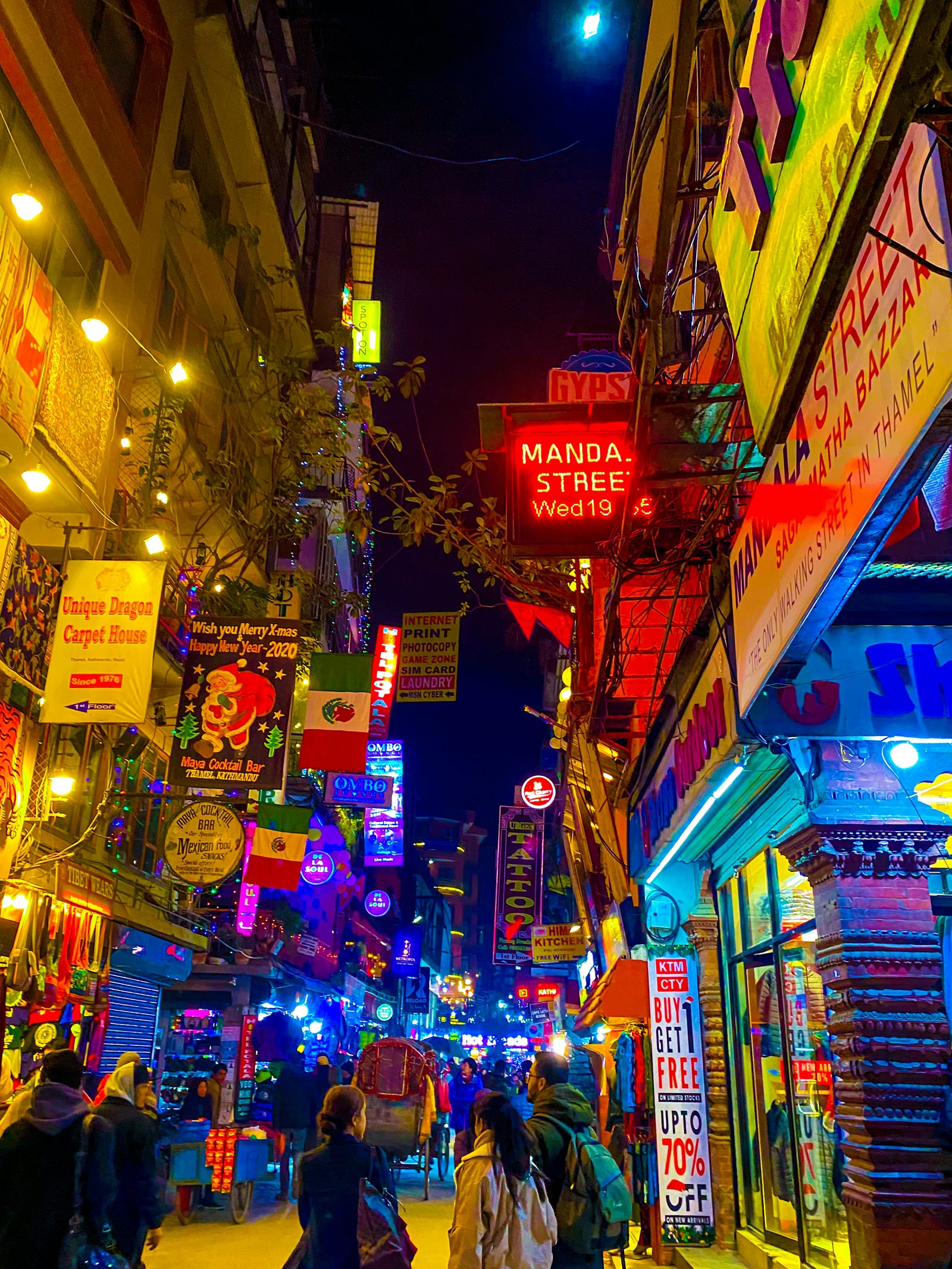 A narrow Thamel street in Kathmandu at night, where Thamel nightlife spills onto the lane, lit by neon signs, string lights, and shopfronts, with walkers heading along the street.