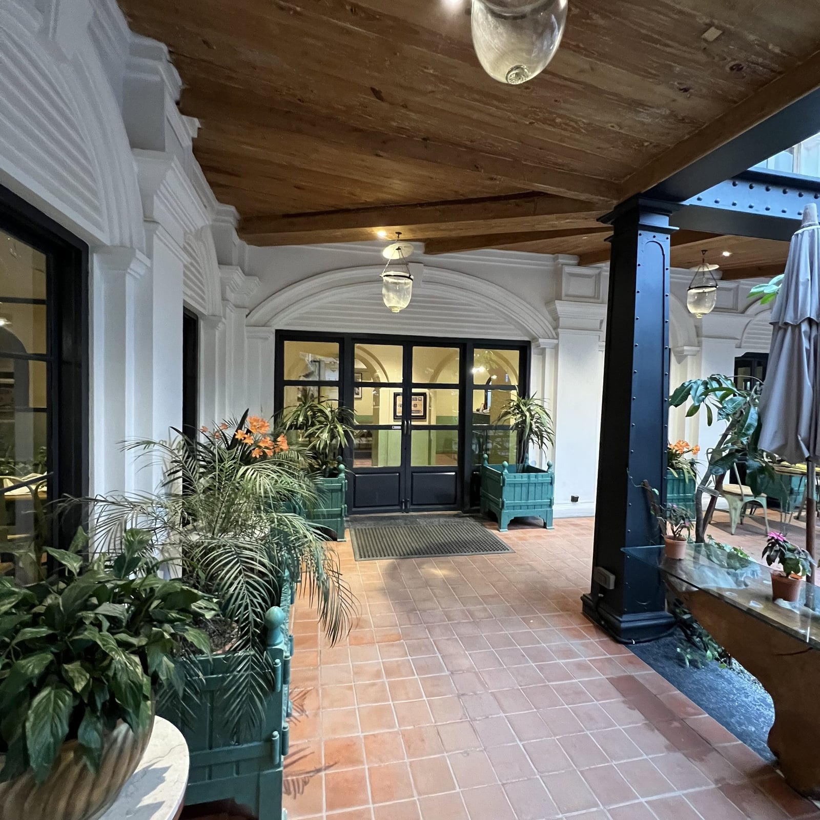The covered arcade entrance walkway of the Neo-Classical Wing at Nepali Ghar Hotel, lined with palms and planter boxes beside the glass entrance doors.