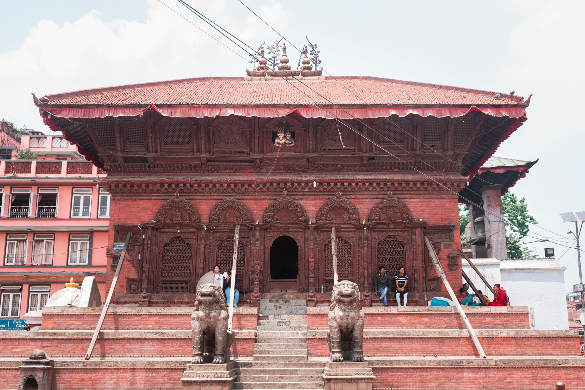 A Newari temple at Kathmandu Durbar Square with a red-brick plinth, intricately carved timber windows, and a pair of stone lions guarding the steps.