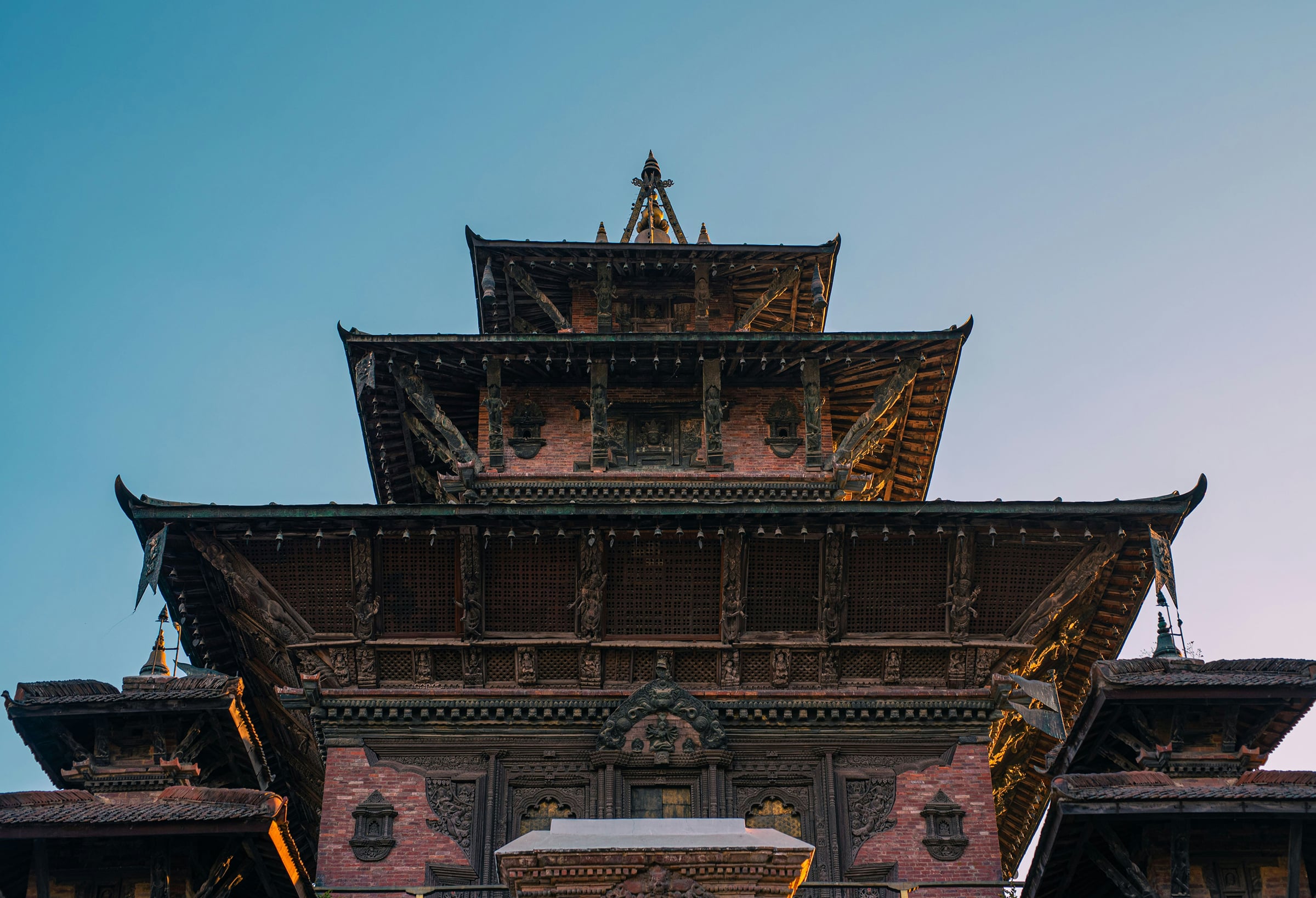 Looking up at a tiered, hand-carved timber pagoda in Kathmandu, with its stacked roofs and carved struts rising against a clear blue evening sky.