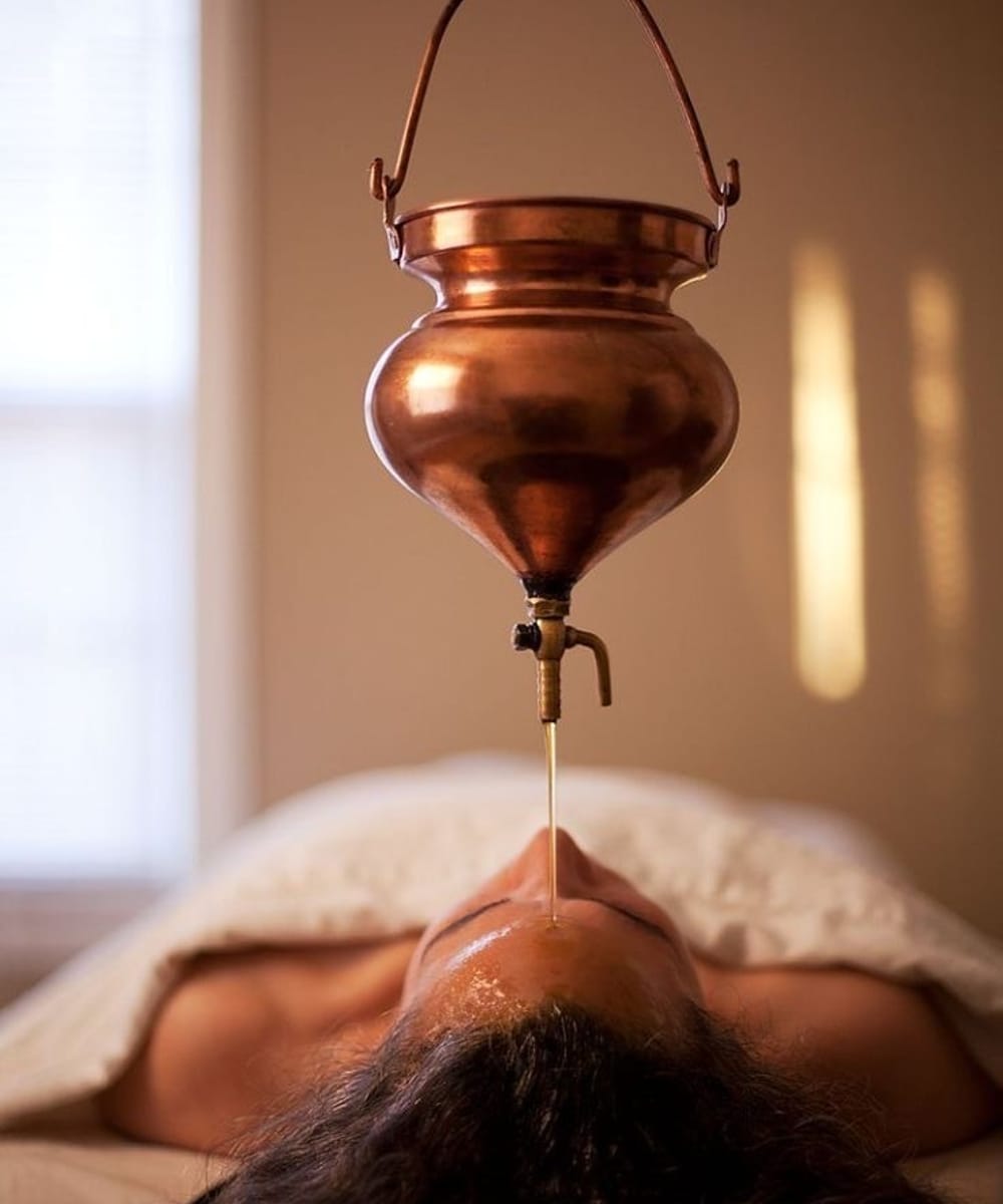 Shirodhara Ayurvedic treatment at Parampara Spa with a copper vessel and brass tap streaming warm oil onto a client's forehead, Sanskreti