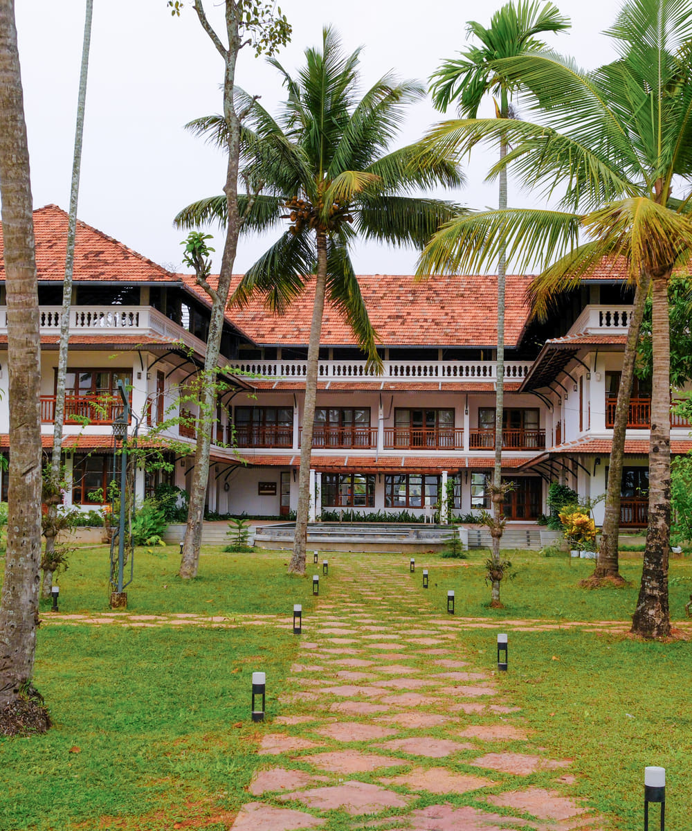 White heritage-style building with red-tiled roof rising above coconut palms and lawn, Sanskreti, Kallanchery Island, Kumbalanghi