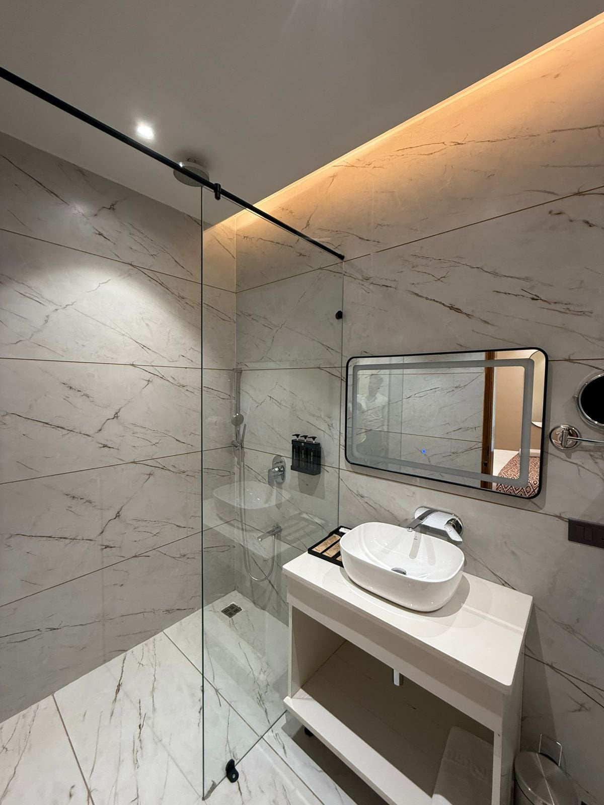 A guest bathroom at Amara Portico, with a walk-in shower behind a glass screen, a washbasin on a counter and a lit mirror