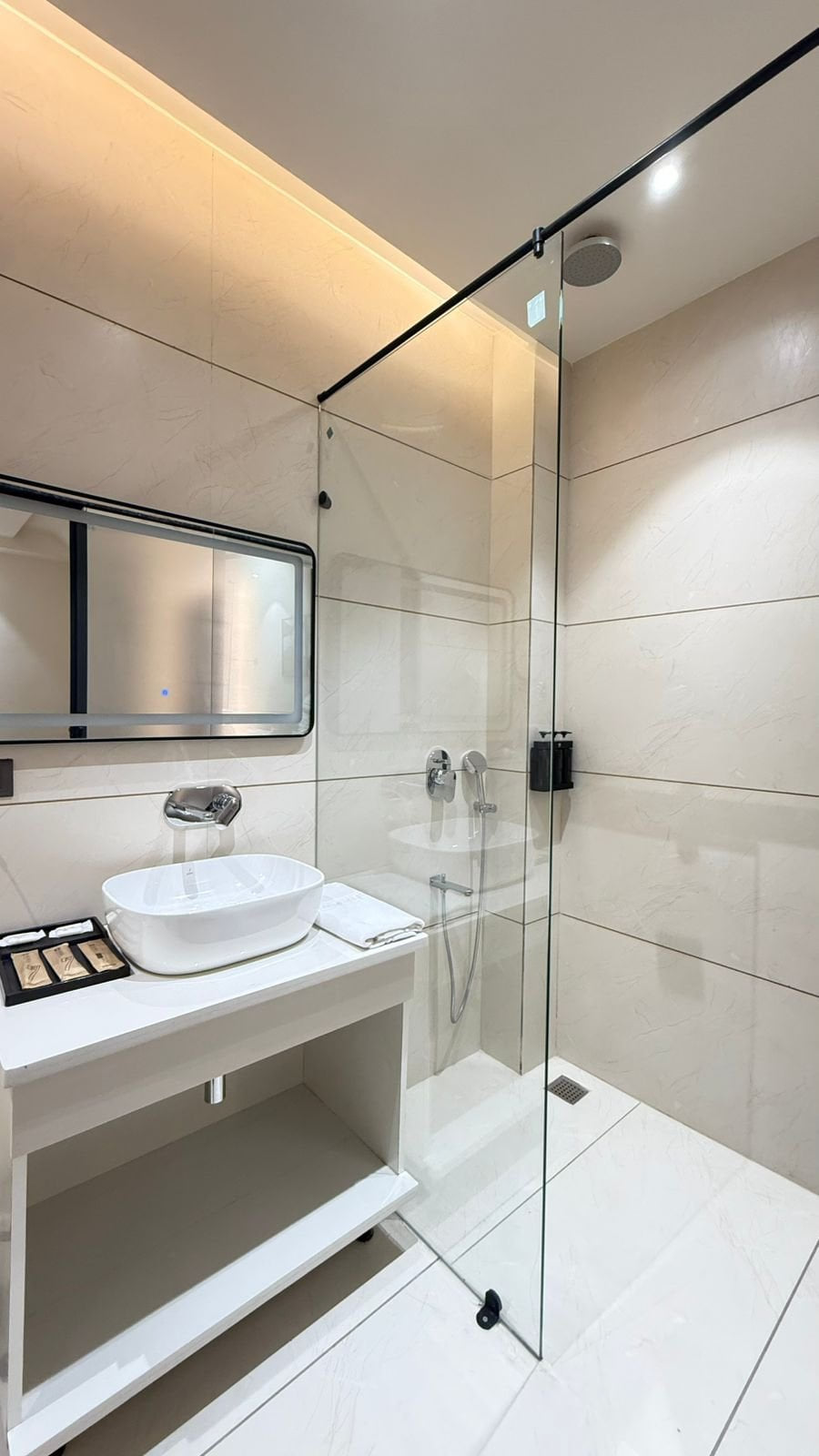 A guest bathroom at Amara Portico, with a glass shower screen, a washbasin set on a counter and a mirror on the tiled wall