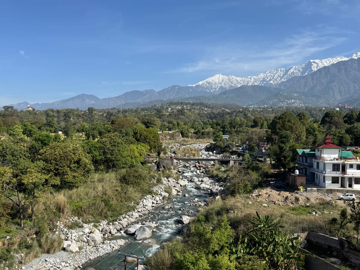The boulder-strewn river valley near Palampur, with houses along the bank and the snow line of the Dhauladhar range beyond
