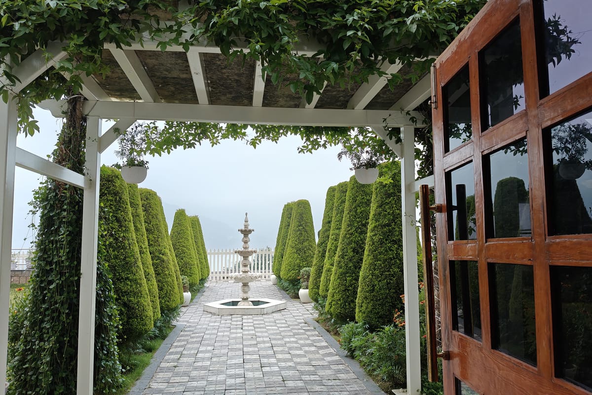 The pergola walk at Levelup The Heritage Kud, clipped conifers lining the paved path down to a stone fountain