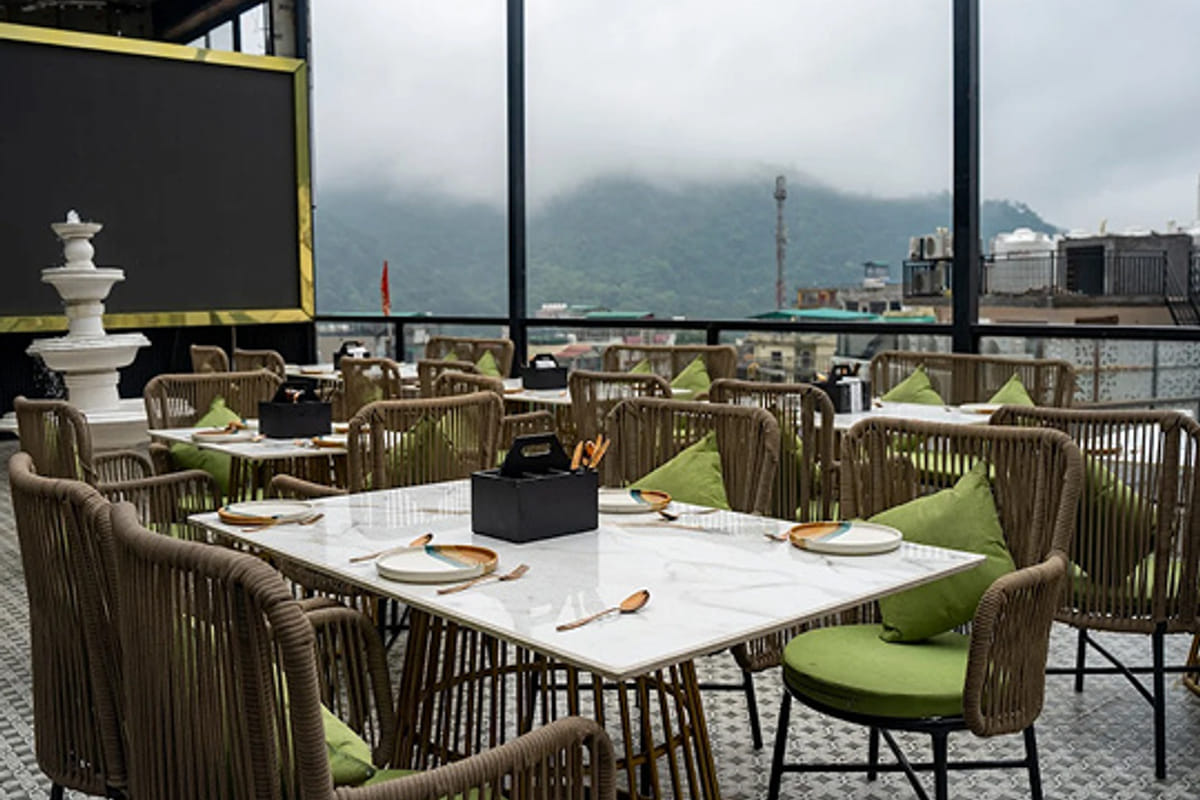 An open-air terrace laid for dining, with cane chairs and green cushions and misty hills rising beyond the railing