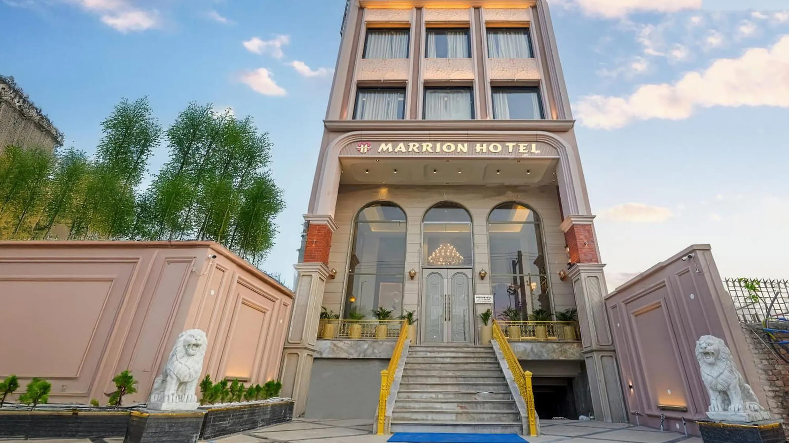 The pale pink facade of Marrion Hotel in Amritsar, with white lion statues either side of the marble steps up to the lit