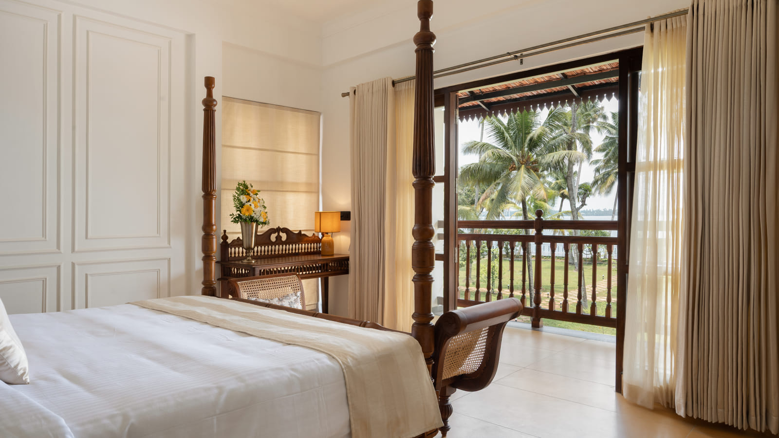 Heritage Suite bedroom with open balcony doors framing coconut palms and the lake, writing desk beside the bed, Sanskreti