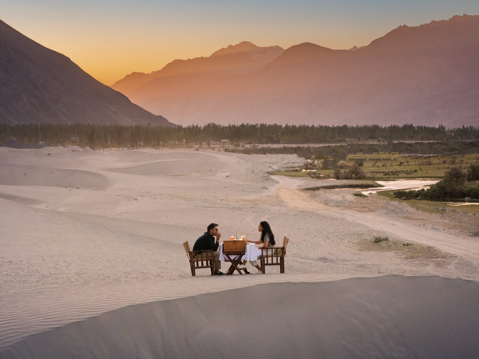 Couple at a sundowner table on the sand dunes against the Karakoram range at twilight