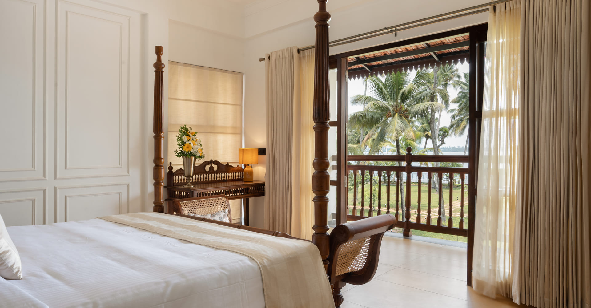 Heritage Suite bedroom with four-poster bed, writing desk and open balcony doors framing coconut palms and the lake, Sanskreti, Kumbalanghi