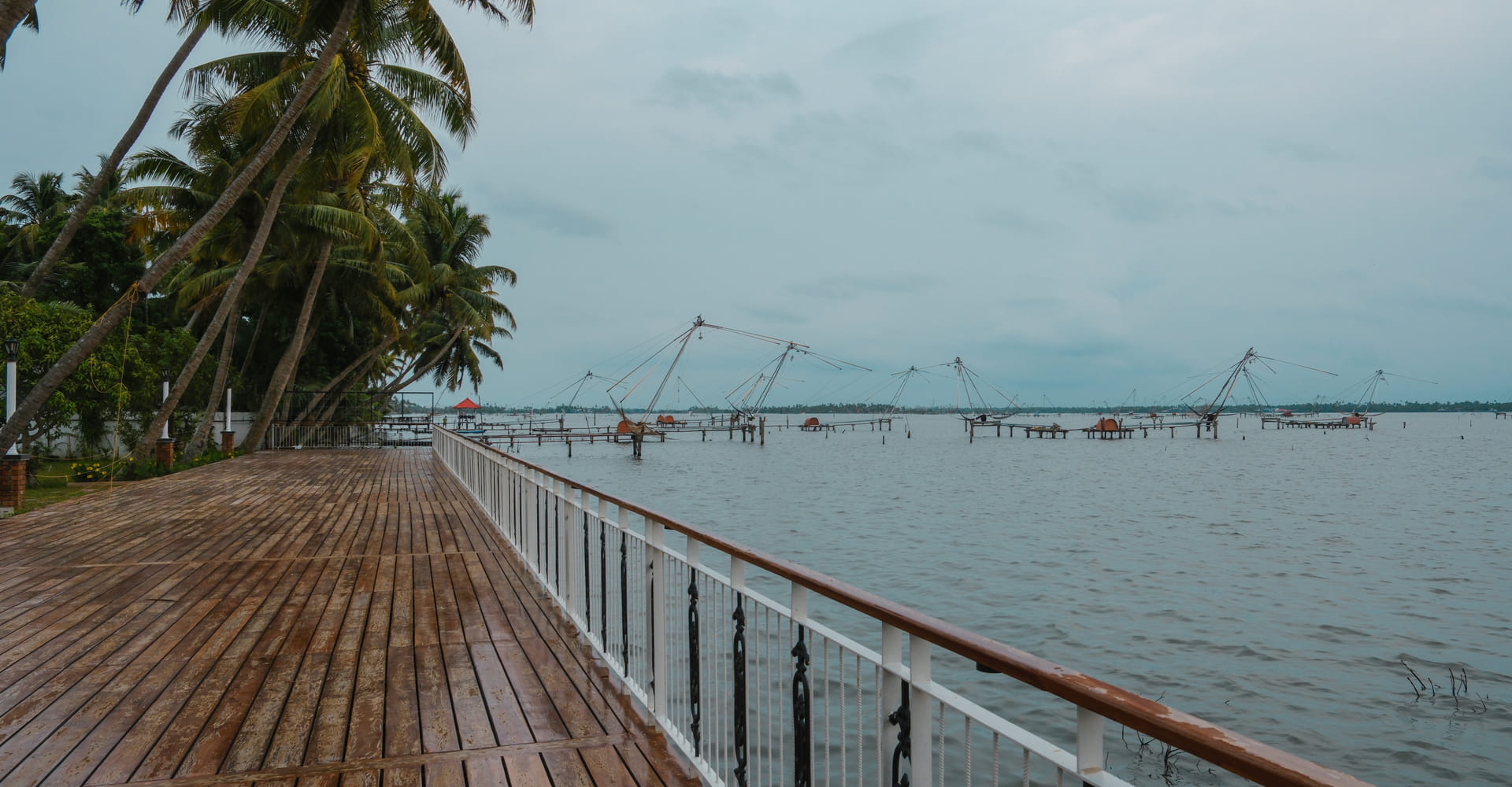 Wooden deck over the backwater with a Chinese fishing net and palm-lined far shore, Sanskreti, Kumbalanghi, Kochi