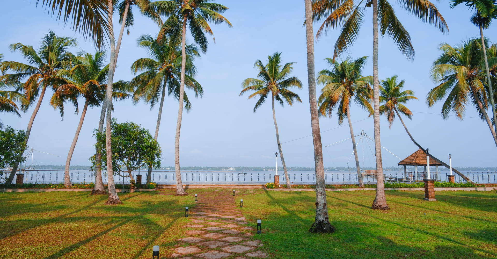 Open event lawn along Vembanad Lake fringed by coconut palms with the palm-lined shoreline behind, Sanskreti