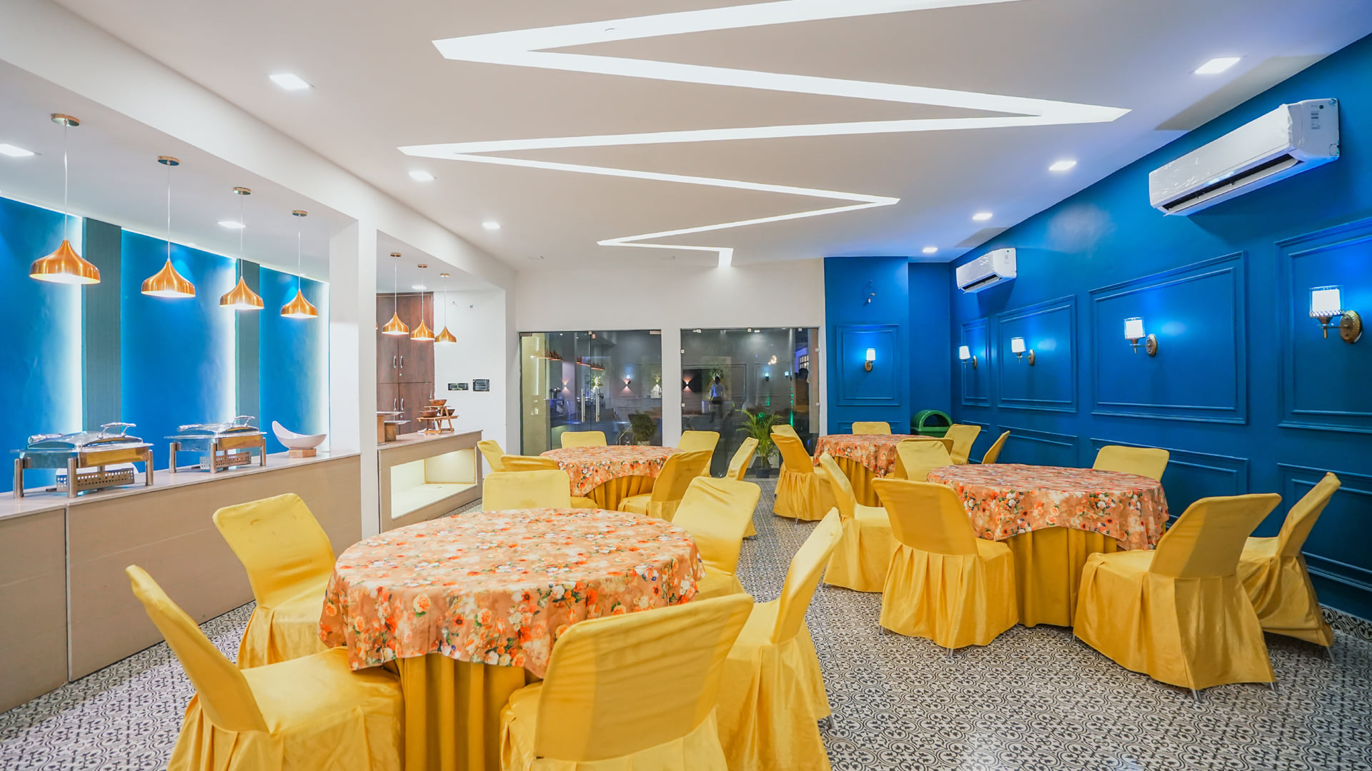 The banquet hall at Escala Hotel, set with round tables, yellow chair covers and floral cloths against a blue lit wall panel