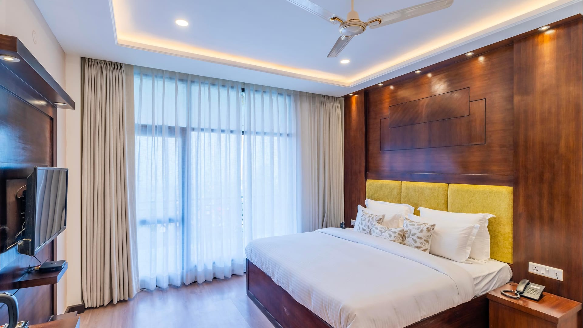A Deluxe room with valley view at Hotel Karnika, with a yellow padded headboard, timber panelling and sheer curtains