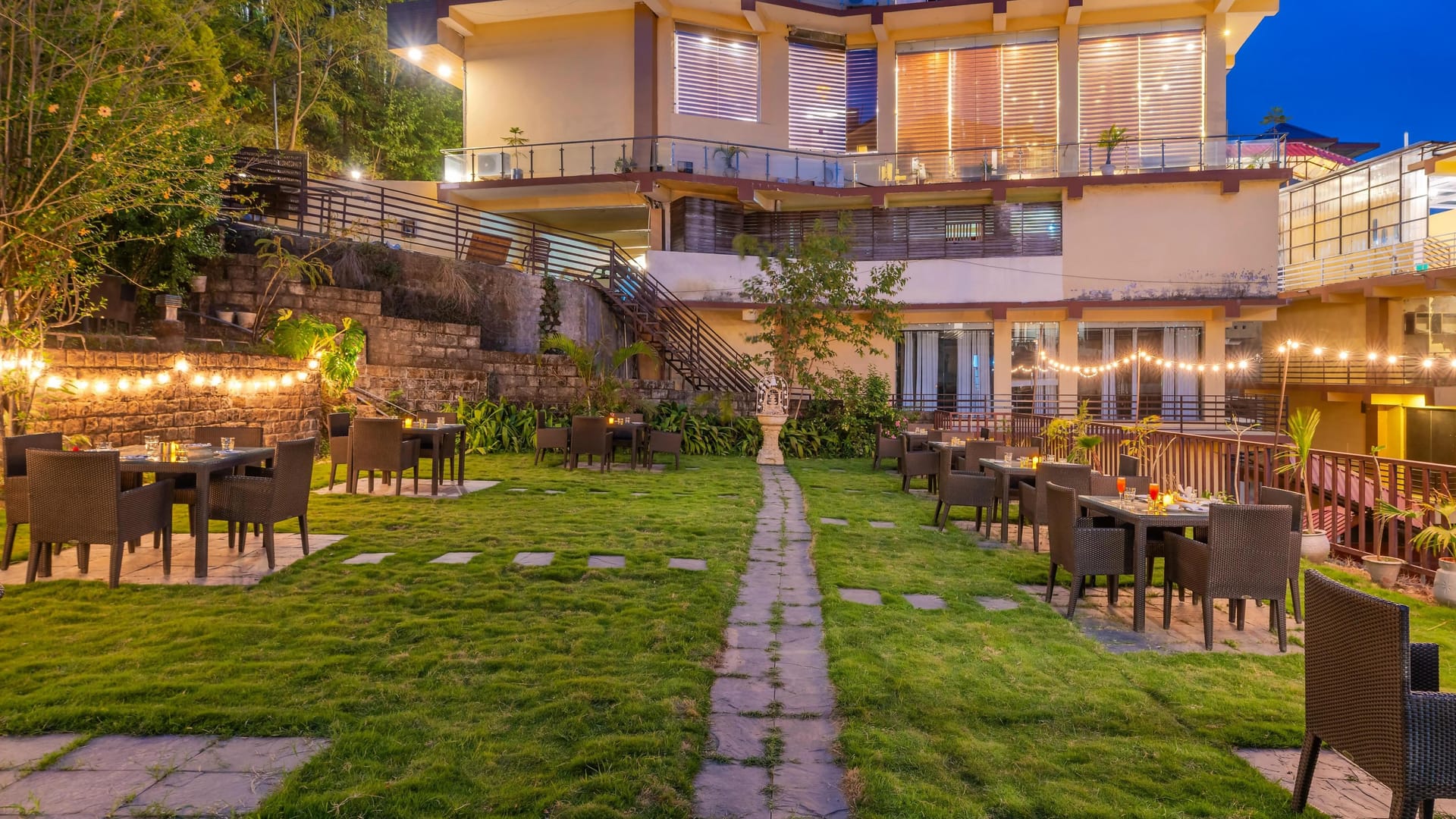The lawn and terrace at Hotel Karnika at dusk, with outdoor dining tables under strings of lights below the guest balconies