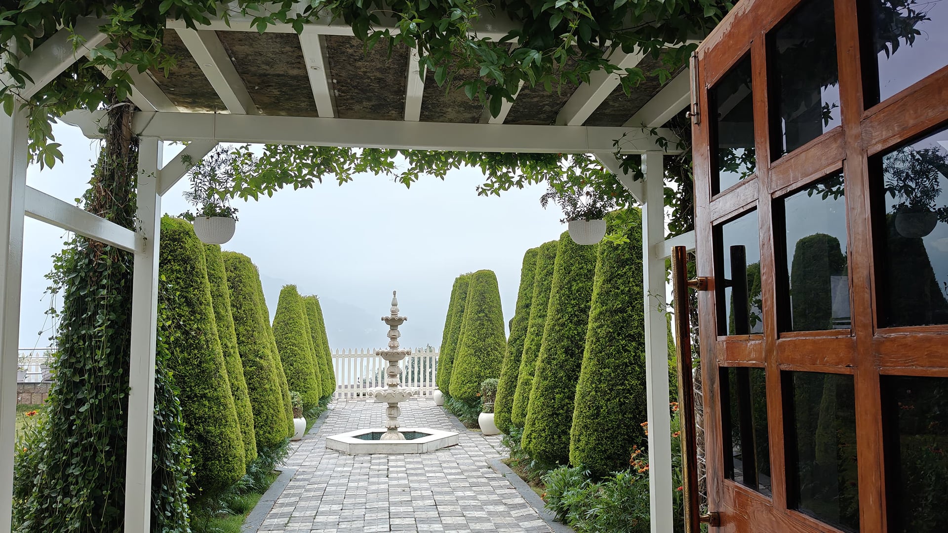 The pergola walk at Levelup The Heritage Kud, clipped conifers lining the paved path down to a stone fountain