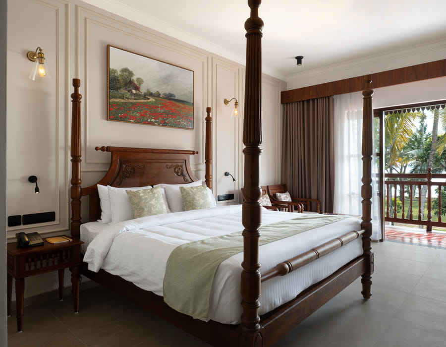 Lake View Room four-poster king bed with cane headboard and balcony doors opening onto palms and Vembanad Lake