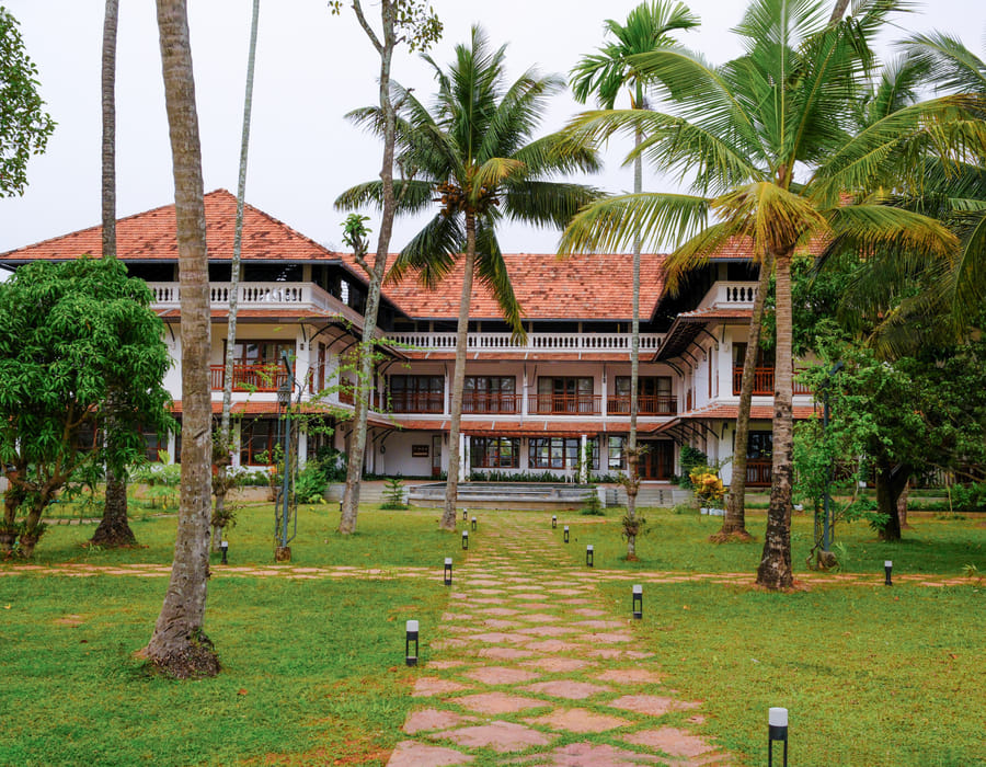 Sanskreti exterior with sloping tiled roof, wooden columns and a landscaped garden along the driveway