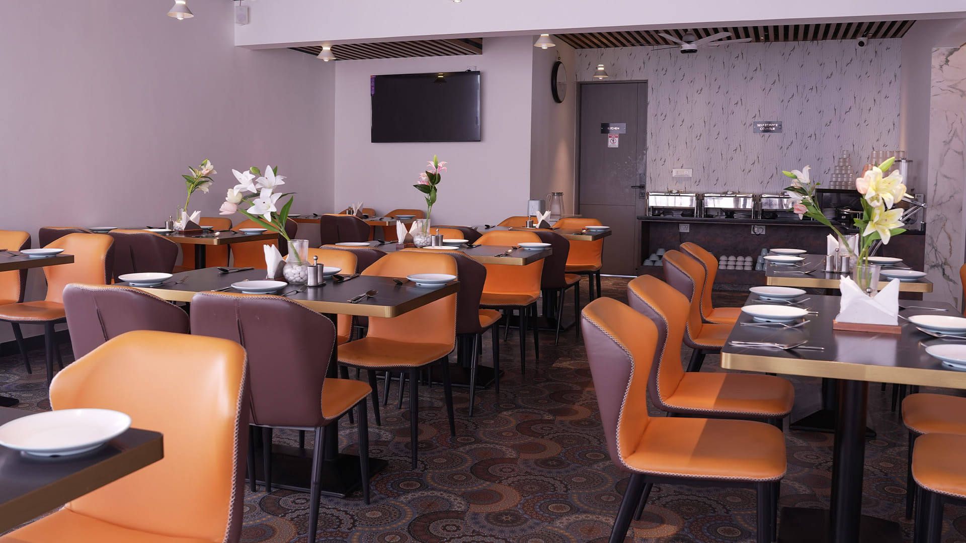 Dining area with orange chairs, a wall-mounted television, and the marble-clad lift lobby beyond