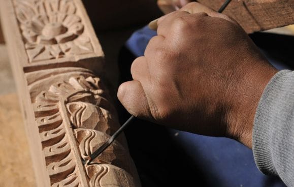 A Newari craftsman's hands chiselling a floral frieze into a timber temple strut at a wood-carving workshop, the same craft seen throughout Nepali Ghar Hotel.