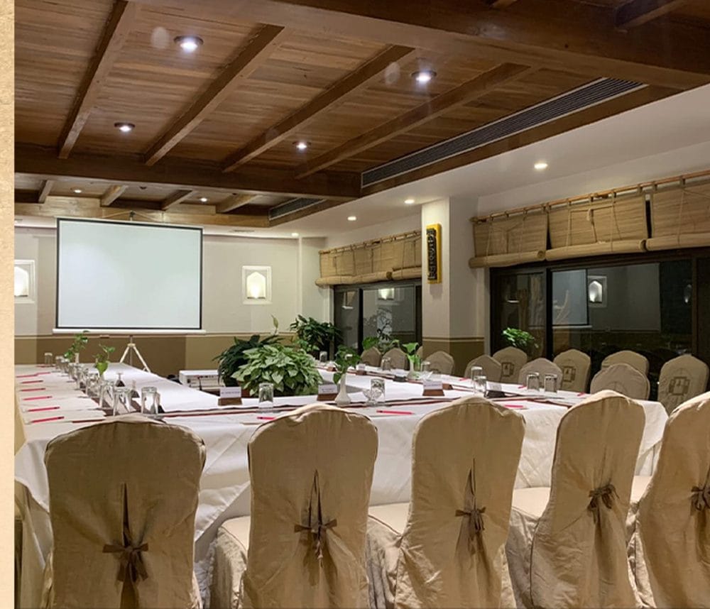 The Baithak meeting room at Nepali Ghar Hotel set up for a conference, with rows of draped chairs facing a lit projector screen beneath a timber-beamed ceiling.