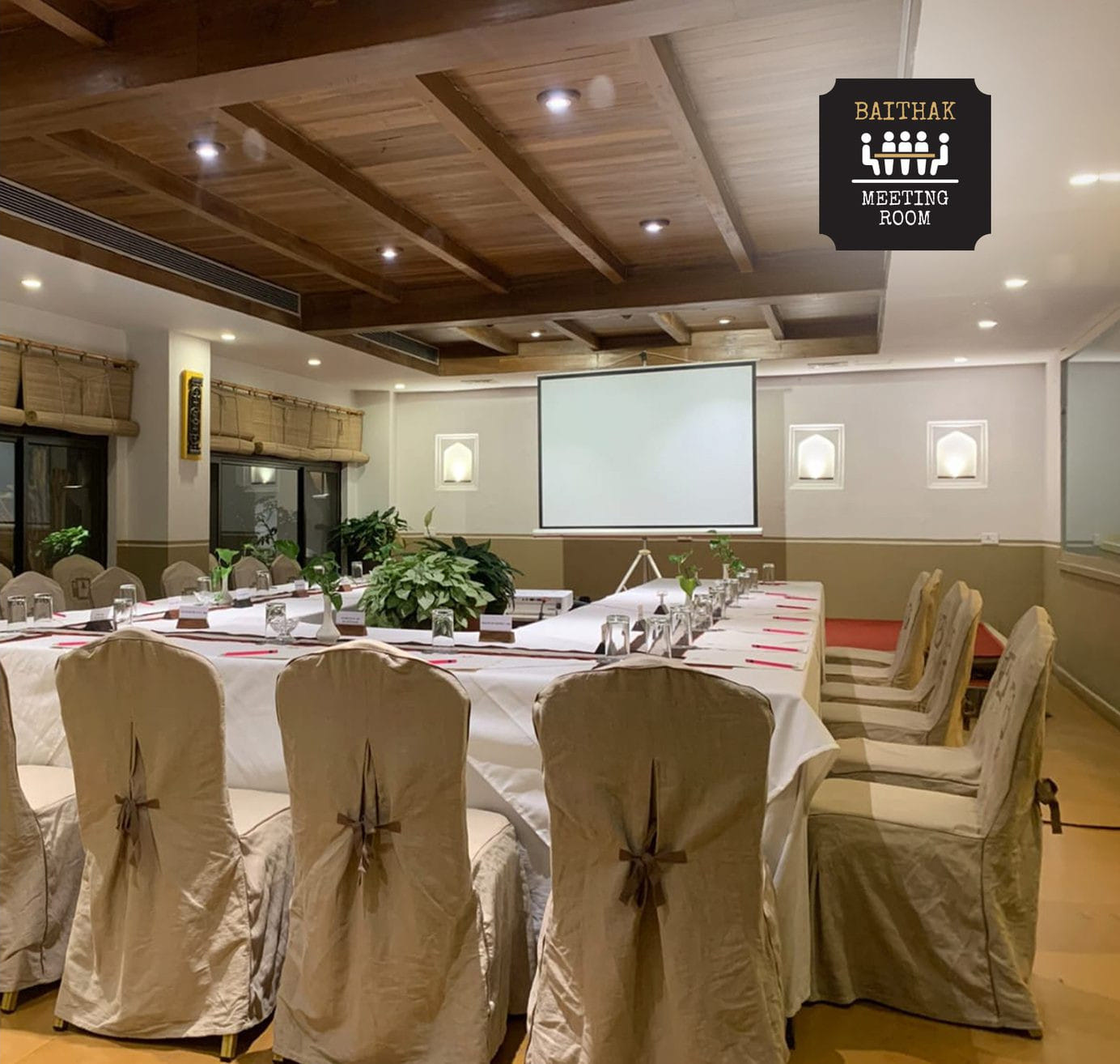 The Baithak meeting room at Nepali Ghar Hotel, arranged in a U-shape setup with white draped chairs around a long table facing a wall-mounted projector screen.