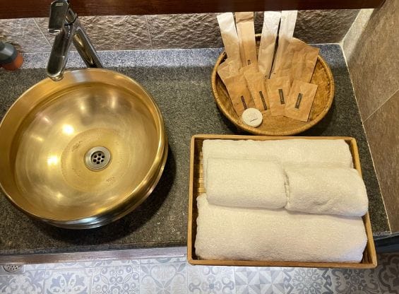 Freshly rolled white towels beside a brass basin on a guest bathroom counter at Nepali Ghar Hotel, part of its towel-and-linen reuse sustainability programme.