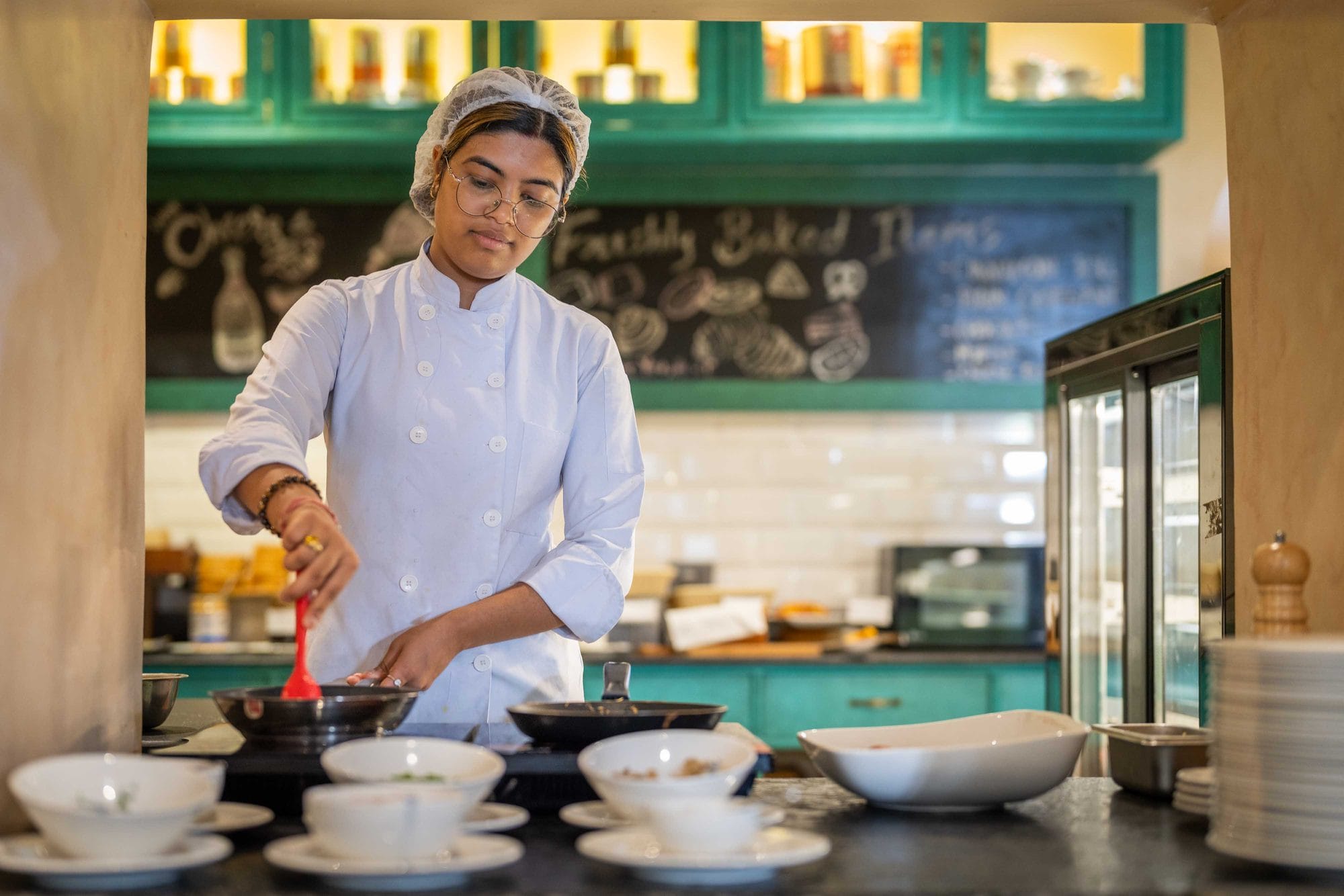 A chef finishing a plated dish at the counter of Chiya Pasal restaurant, Nepali Ghar Hotel, beneath the hand-chalked menu board listing the day's specials.