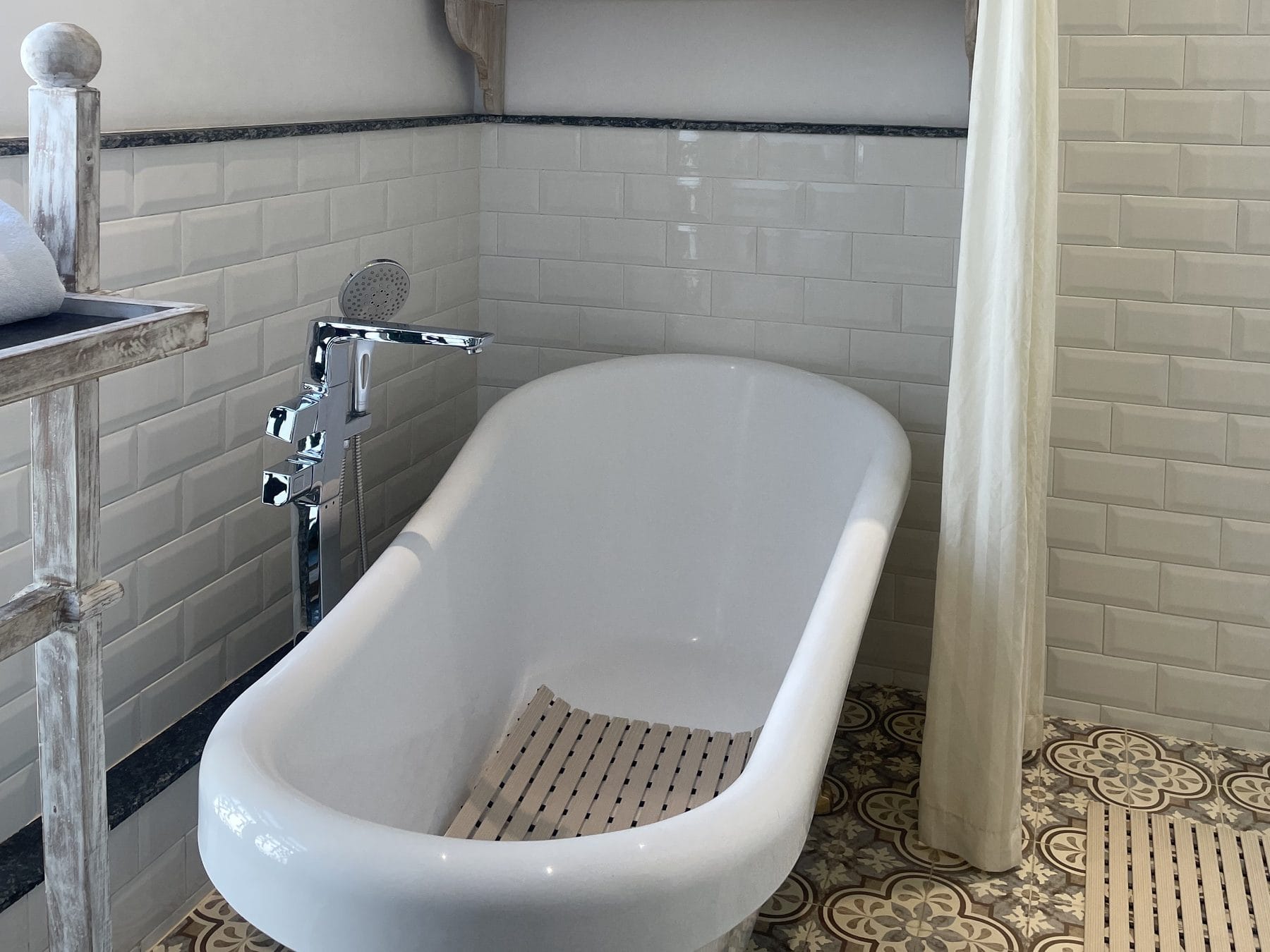 A clawfoot bathtub in the Classic Junior Suite bathroom at Nepali Ghar Hotel, set on a patterned tile floor beside a wooden shelf holding fresh white towels.