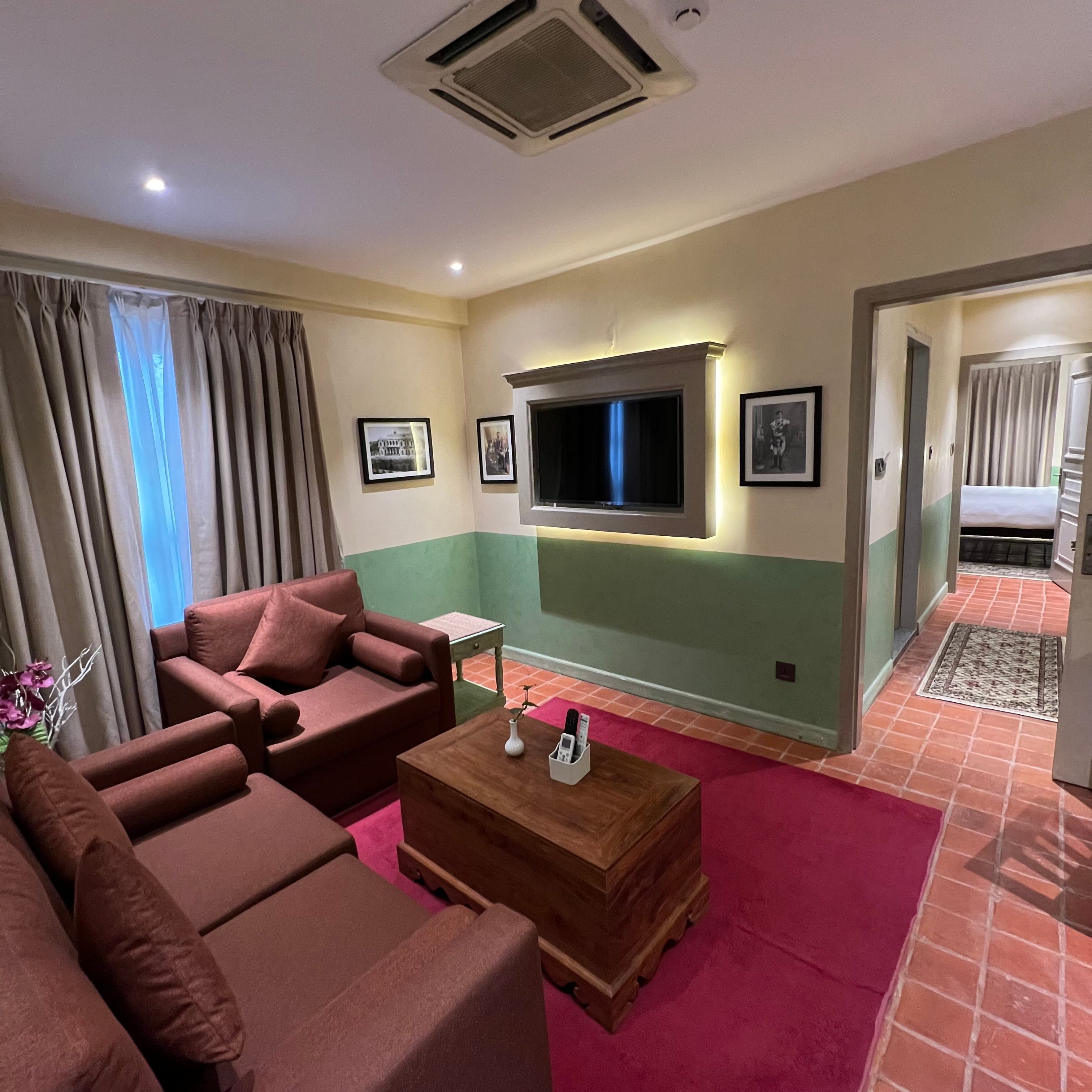 The separate living room of the Classic Junior Suite at Nepali Ghar Hotel, with a wine-red sofa set, a wall-mounted television, and a doorway through to the bedroom.