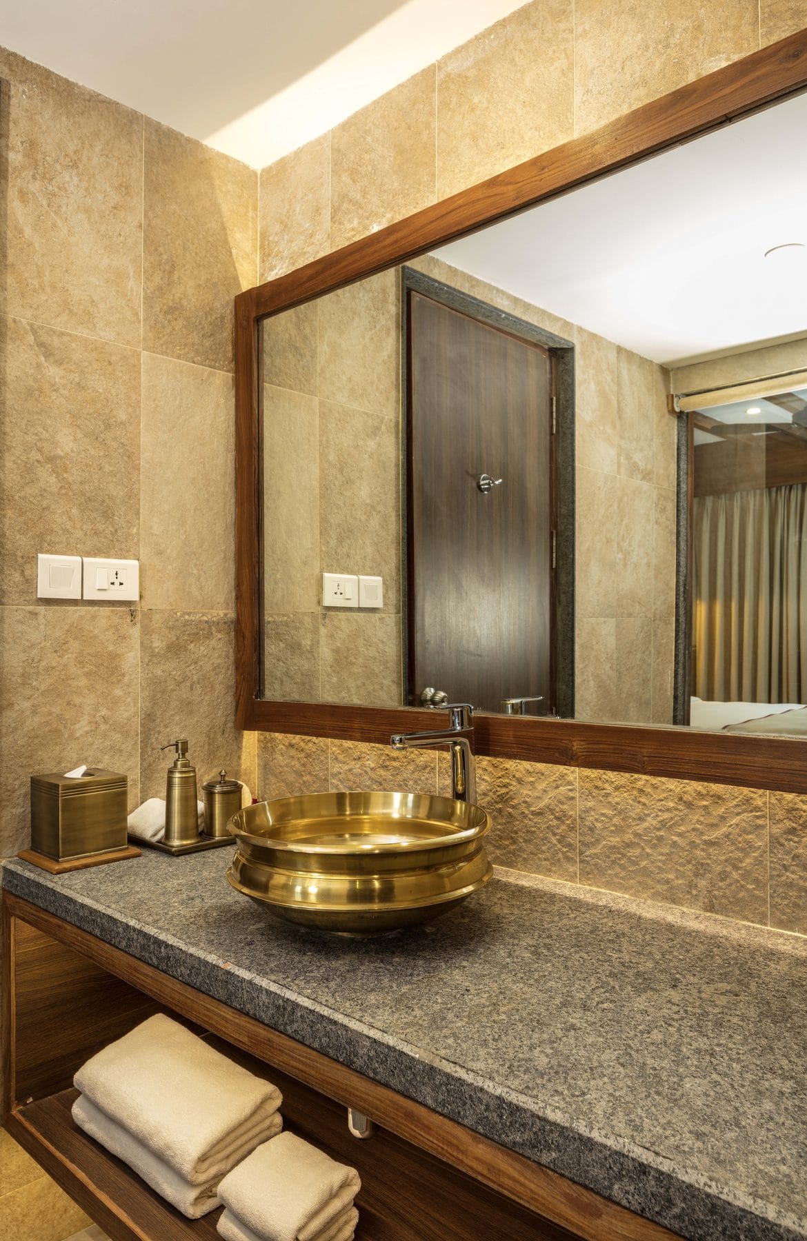 The Executive Suite bathroom vanity at Nepali Ghar Hotel, a brass basin set on a stone counter beneath a timber-framed mirror and a row of amenity bottles.