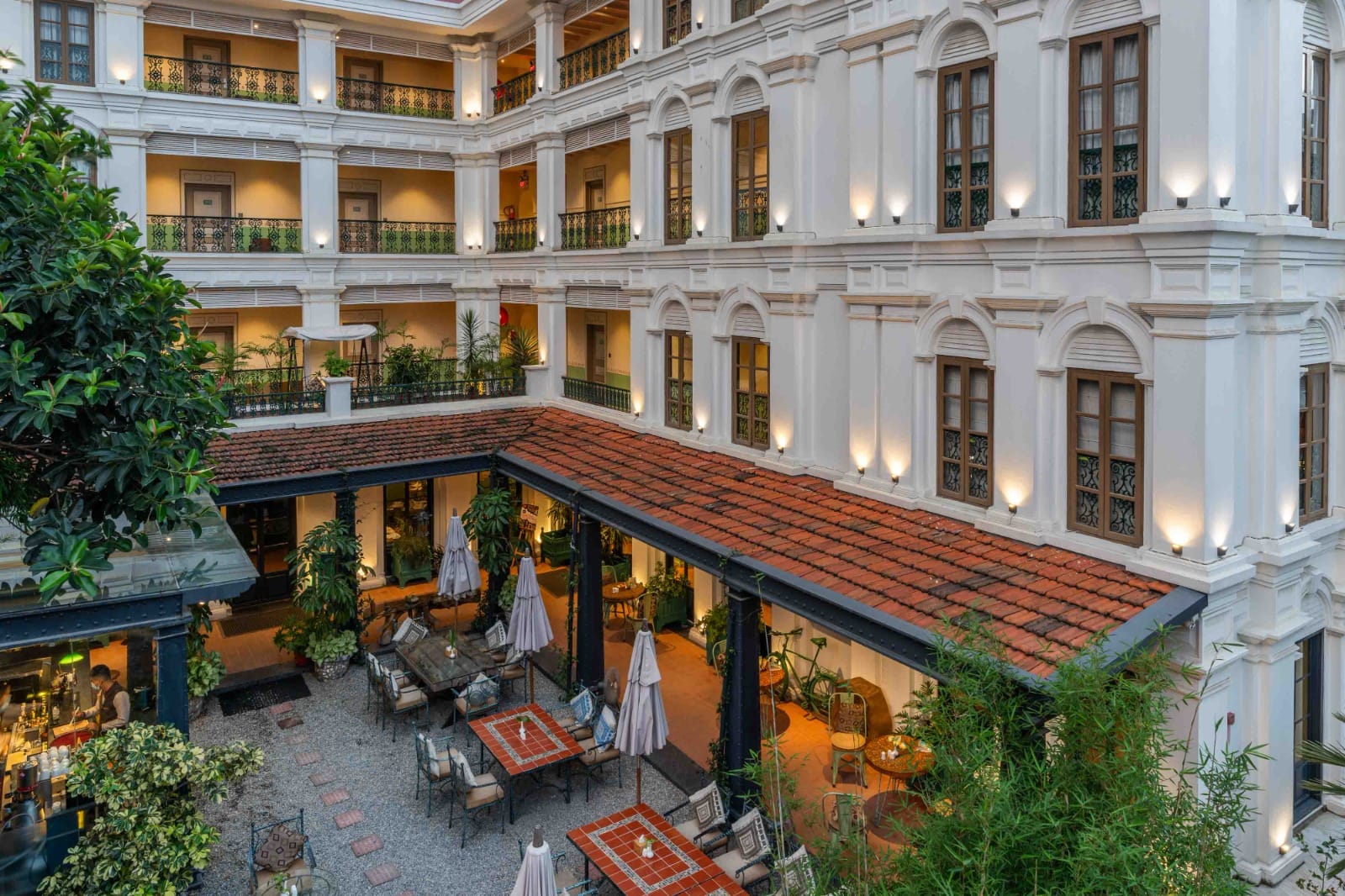 An aerial view at dusk of the planted front yard at Nepali Ghar Hotel, its lush tiled colonnade and parasol-shaded seating ringed by tiered colonial balconies.