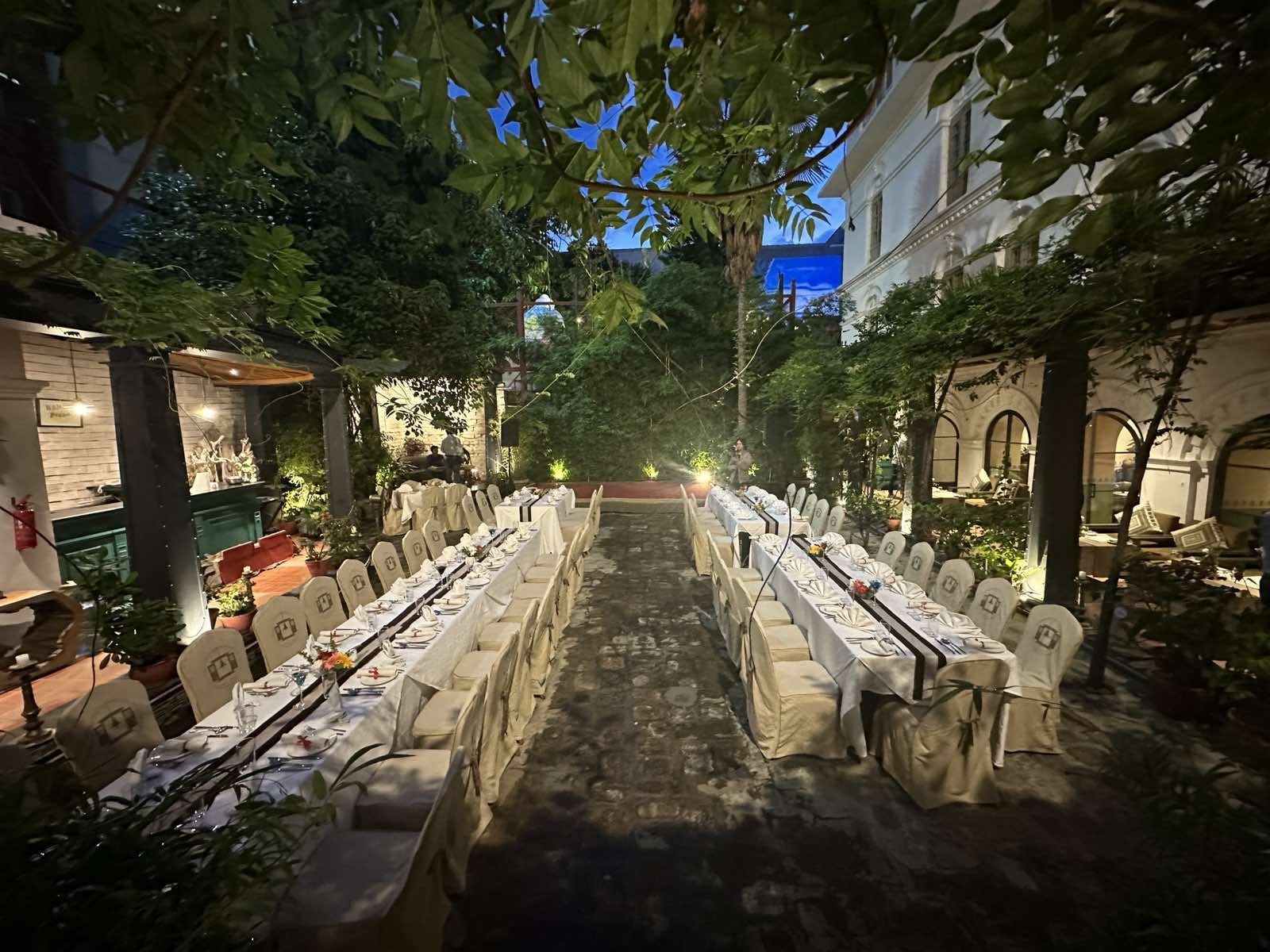Lush greenery bordering the front yard garden at Nepali Ghar Hotel at night, golden string lights strung between the trees above the wrought-iron seating below.