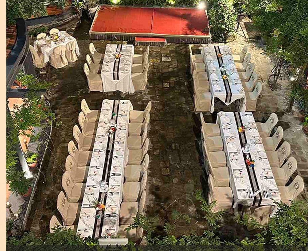 An aerial view of long banquet tables dressed in white linen and set for dinner in the garden courtyard at Nepali Ghar Hotel, beneath a large red event awning.