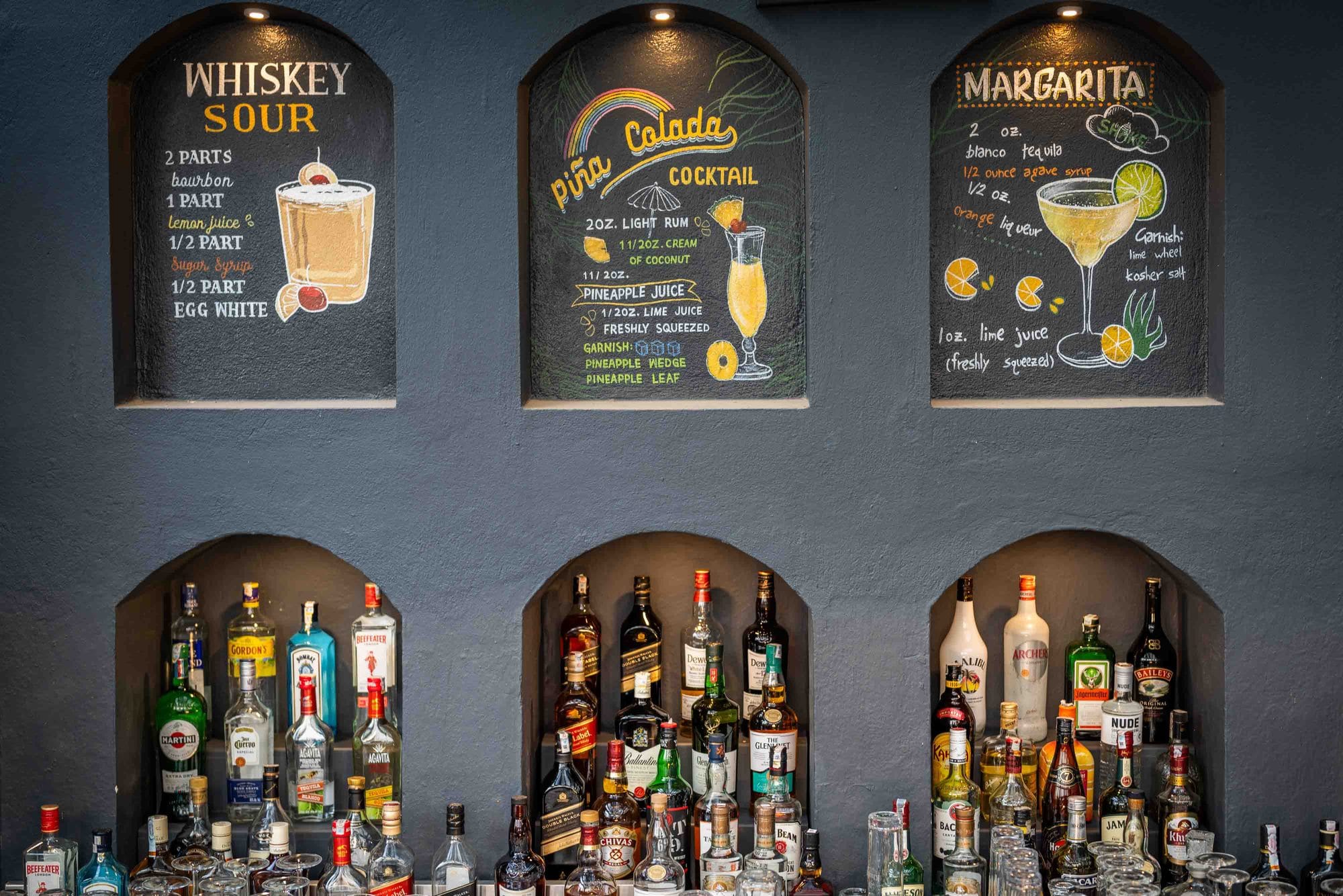 Hand-chalked recipes for a whiskey sour, pina colada, and margarita displayed above the lit spirits niches at Jharana Bar, the outdoor bar of Nepali Ghar Hotel.