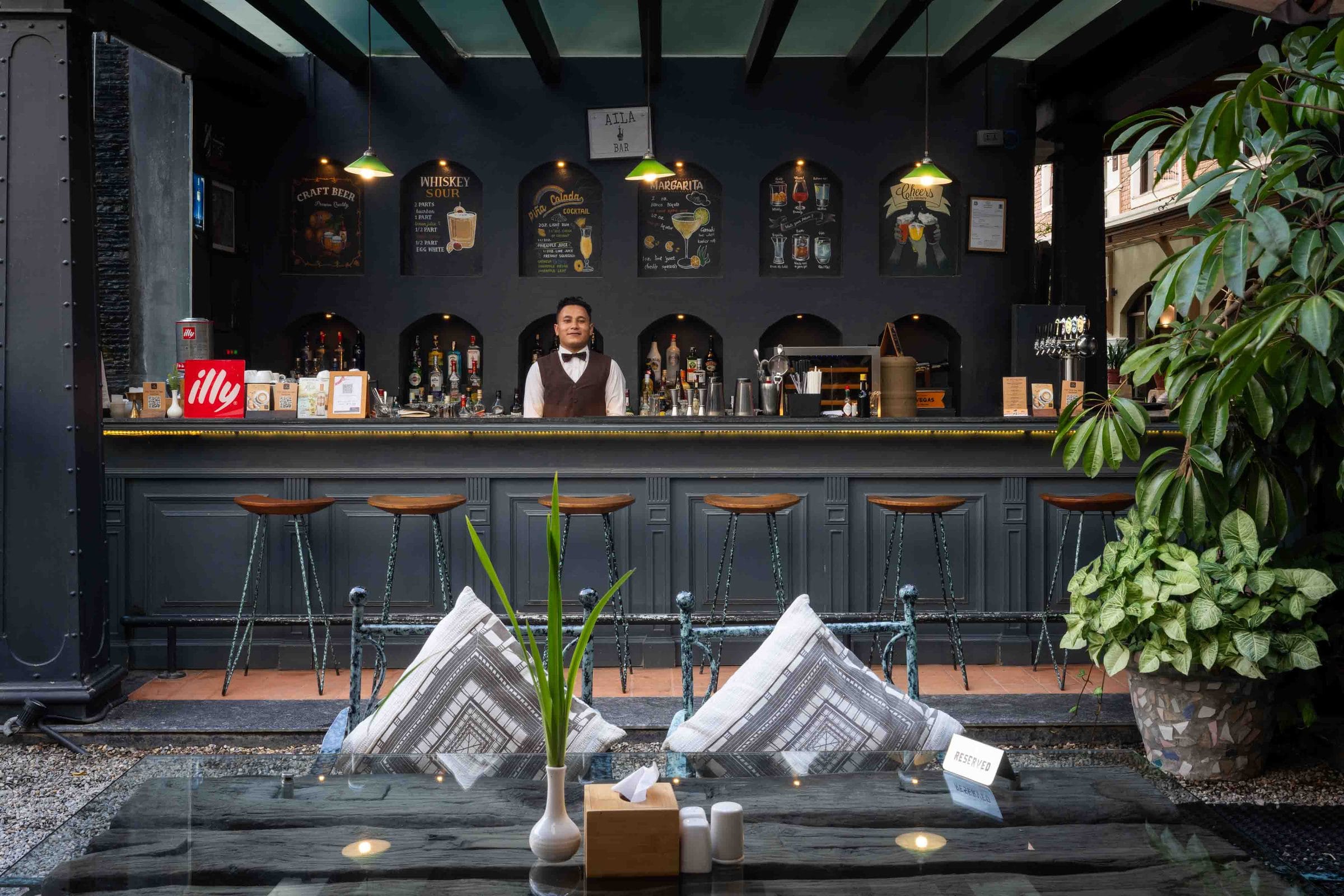 The Jharana Bar counter at Nepali Ghar Hotel at dusk, a bartender behind the taps beneath timber stools and hand-chalked cocktail recipes above the bar itself.
