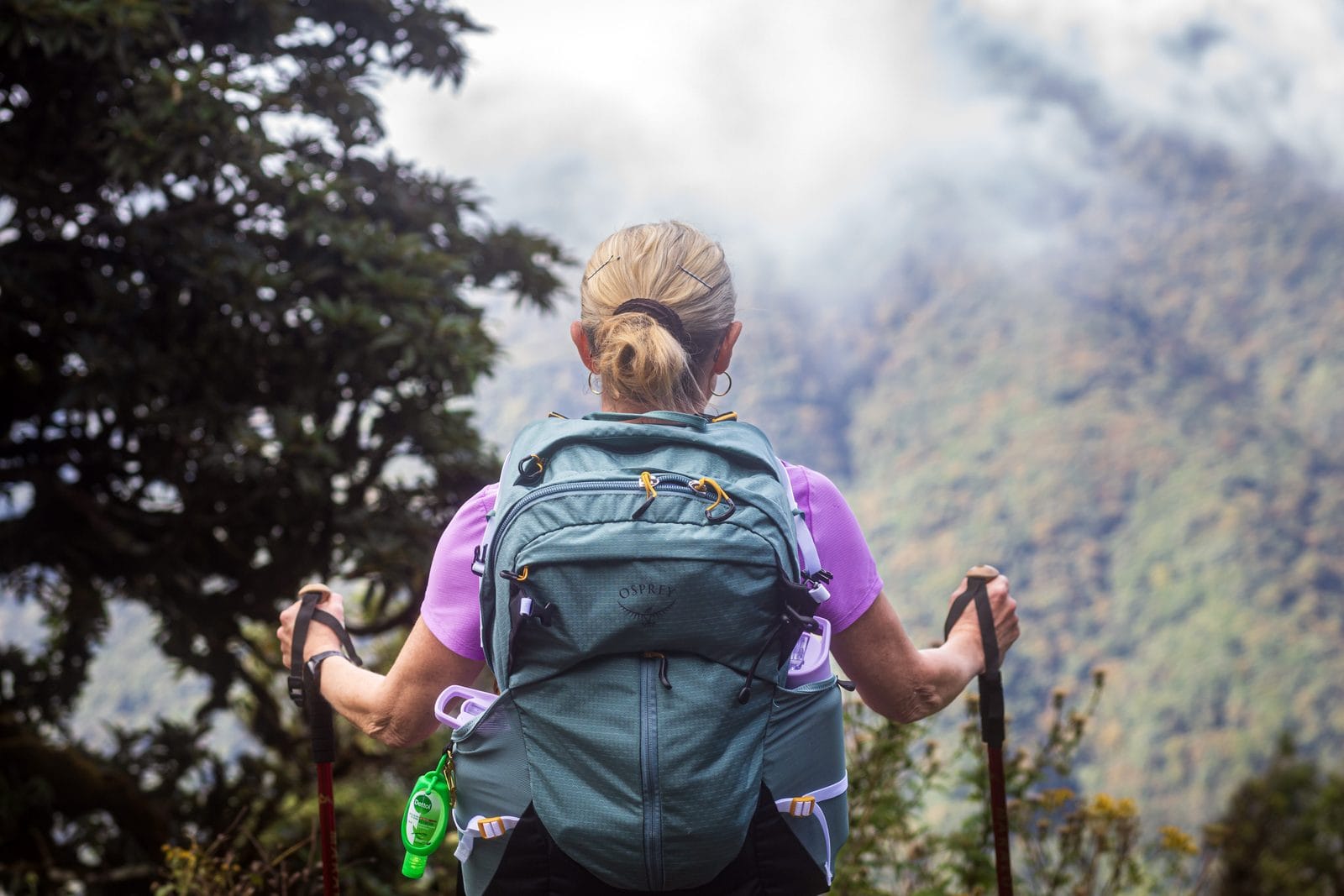 A trekker with walking poles pausing on a forest trail as cloud rolls across the ridge above Kathmandu Valley, one of the guided hikes from Nepali Ghar Hotel.
