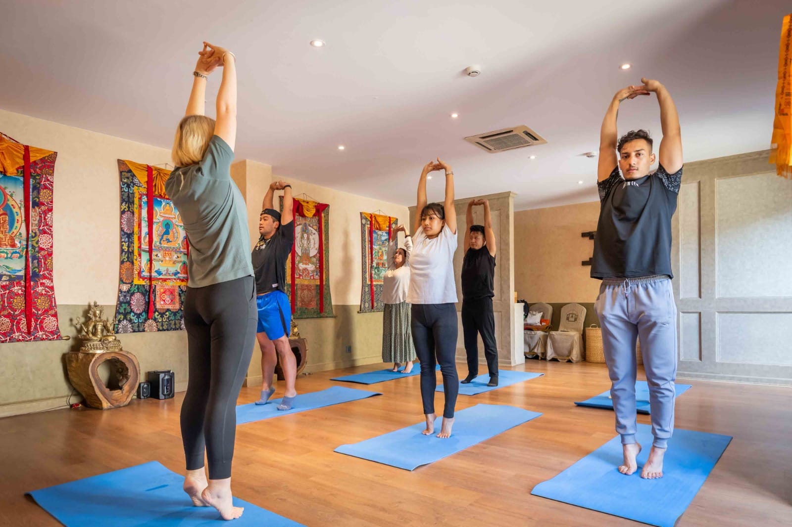 Guests holding a standing overhead stretch during the complimentary yoga class at Nepali Ghar Hotel, framed by thangka scrolls and a small brass Buddha statue.