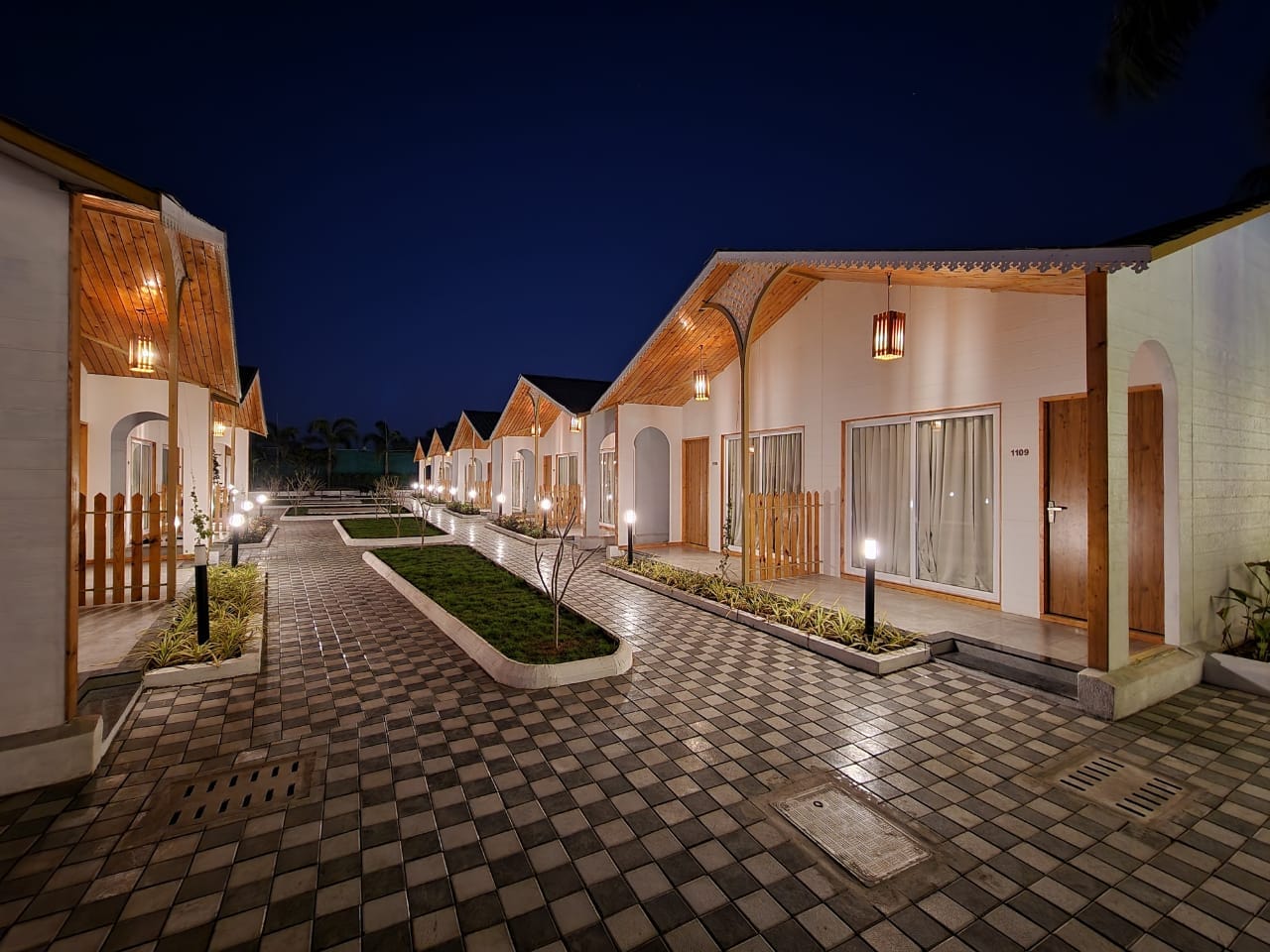 A row of lantern-lit cottages with pitched wooden roofs along a paved pathway at night at VITS Daman Devka Beach