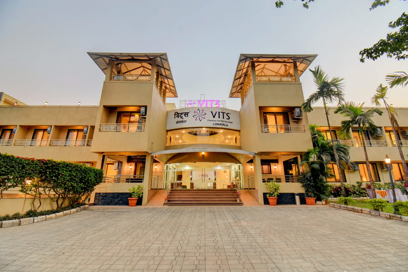 Illuminated entrance and twin glass towers of VITS Lonavala at dusk, with palm trees and a paved forecourt